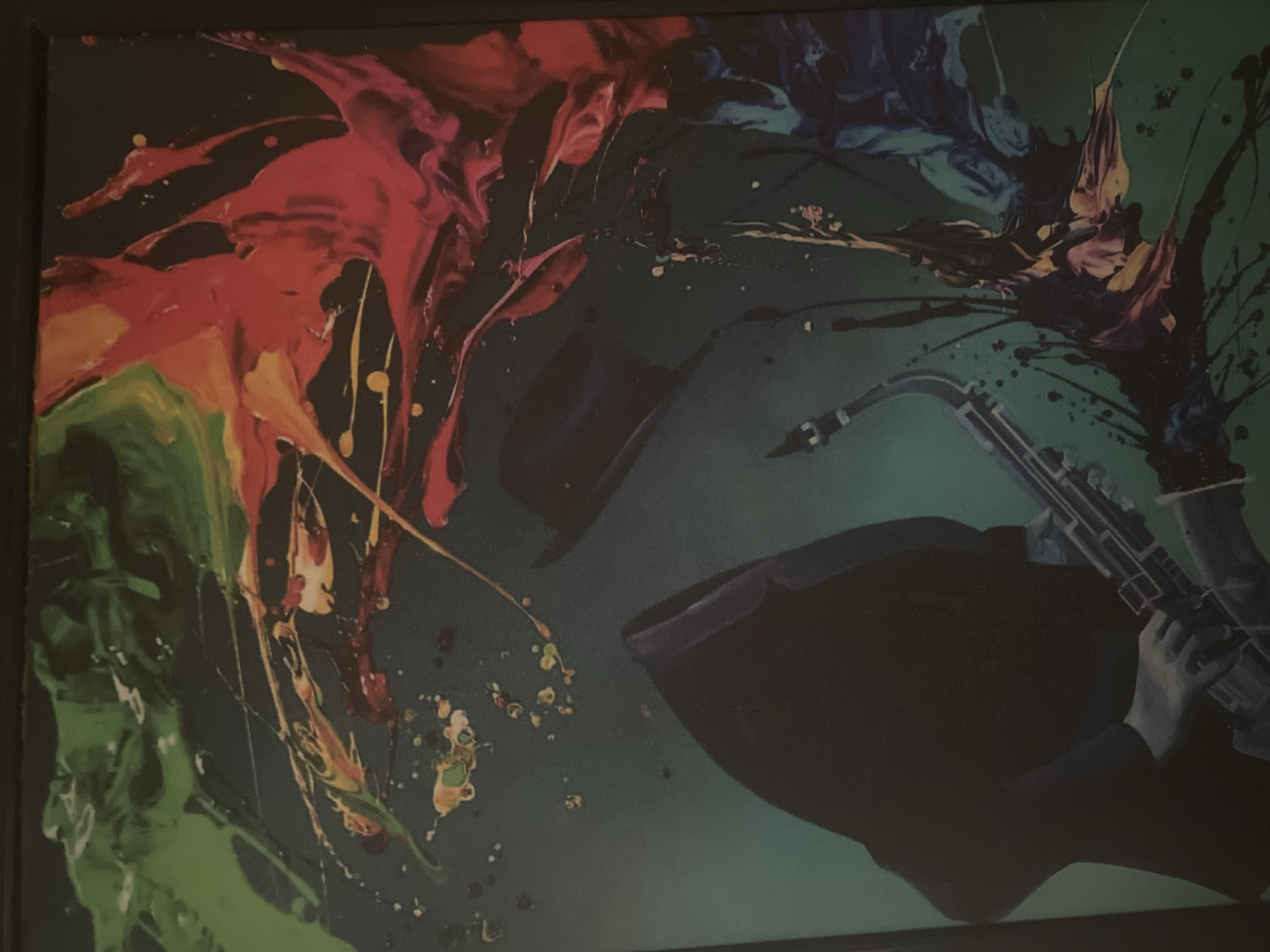 The image presents a dynamic and colorful abstract painting on a dark teal or green background. The artwork features vibrant splashes and drips of paint in various hues, including bright red, orange, green, yellow, and blue, primarily emanating from the top left and top right corners of the canvas, spreading towards the center.
Key subjects in the painting include a dark fedora hat, which appears to be floating in the upper-middle section, surrounded by the colorful paint. In the lower right quadrant, the silhouette of a person's upper body and arms is visible, holding a saxophone. The hands are depicted in a lighter tone, realistically gripping the instrument, suggesting an active performance. The saxophone itself is rendered with some detail, standing out against the dark silhouette of the musician.
The activity depicted is an implied musical performance, likely jazz or blues, given the saxophone and fedora. The energetic paint splatters convey a sense of sound, movement, and emotion, as if the music is visually bursting forth from the instrument and performer. The contrast between the bright, flowing colors and the dark, silhouetted figures creates a lively and expressive mood. There is no visible text in the image.