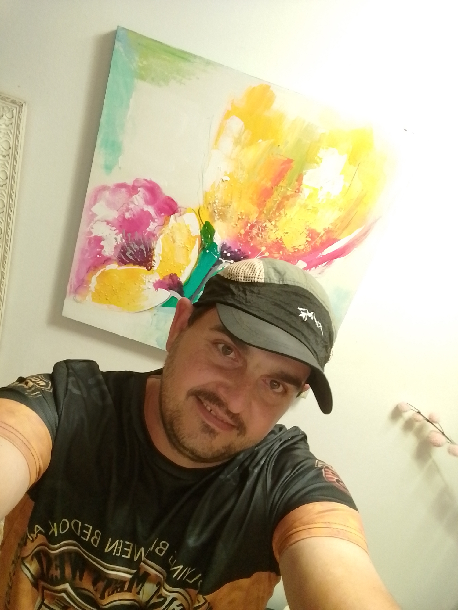 In Wedel, Germany, a man is captured in a selfie. He is wearing a black t-shirt with orange accents and a baseball cap. In the background, a painting with vibrant abstract floral designs is displayed on a white wall. The lighting suggests an indoor setting, possibly a home or studio.