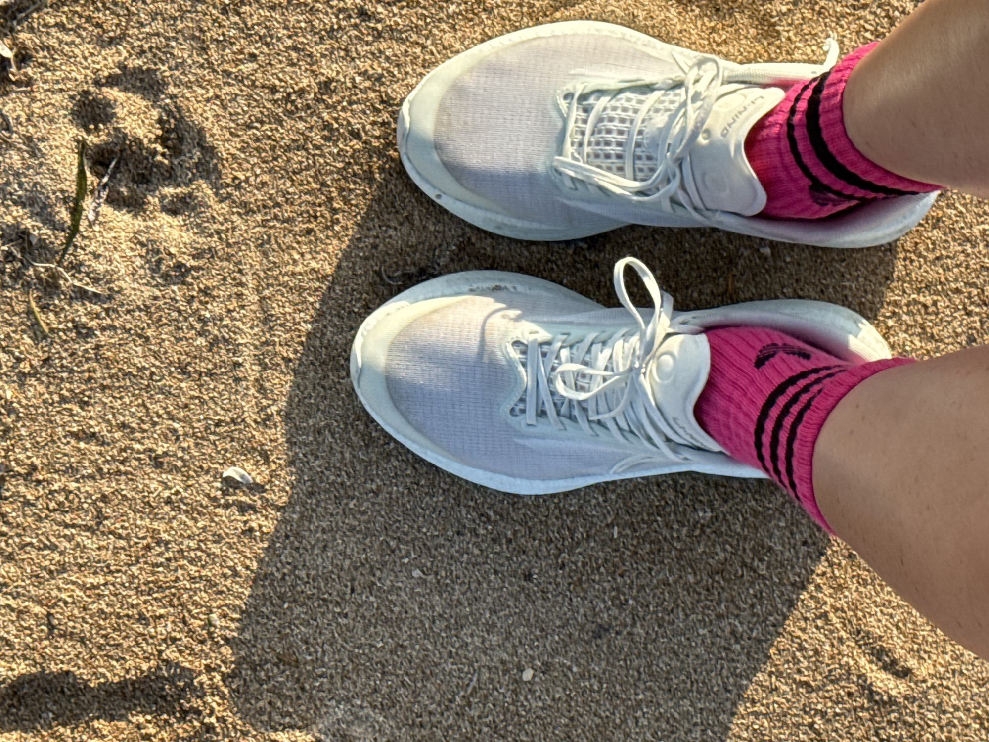 A pair of light blue athletic shoes with white laces are worn by an unseen individual standing on a sandy surface. The person is wearing bright pink ankle socks with three black stripes around the cuff. The sand appears to be on a beach, with a shadow cast from the person's leg and body on the left side of the frame. There are a few blades of dried vegetation and a small white feather or shell fragment on the sand. The image is taken from a top-down perspective. The location is identified as Denpasar, Indonesia.