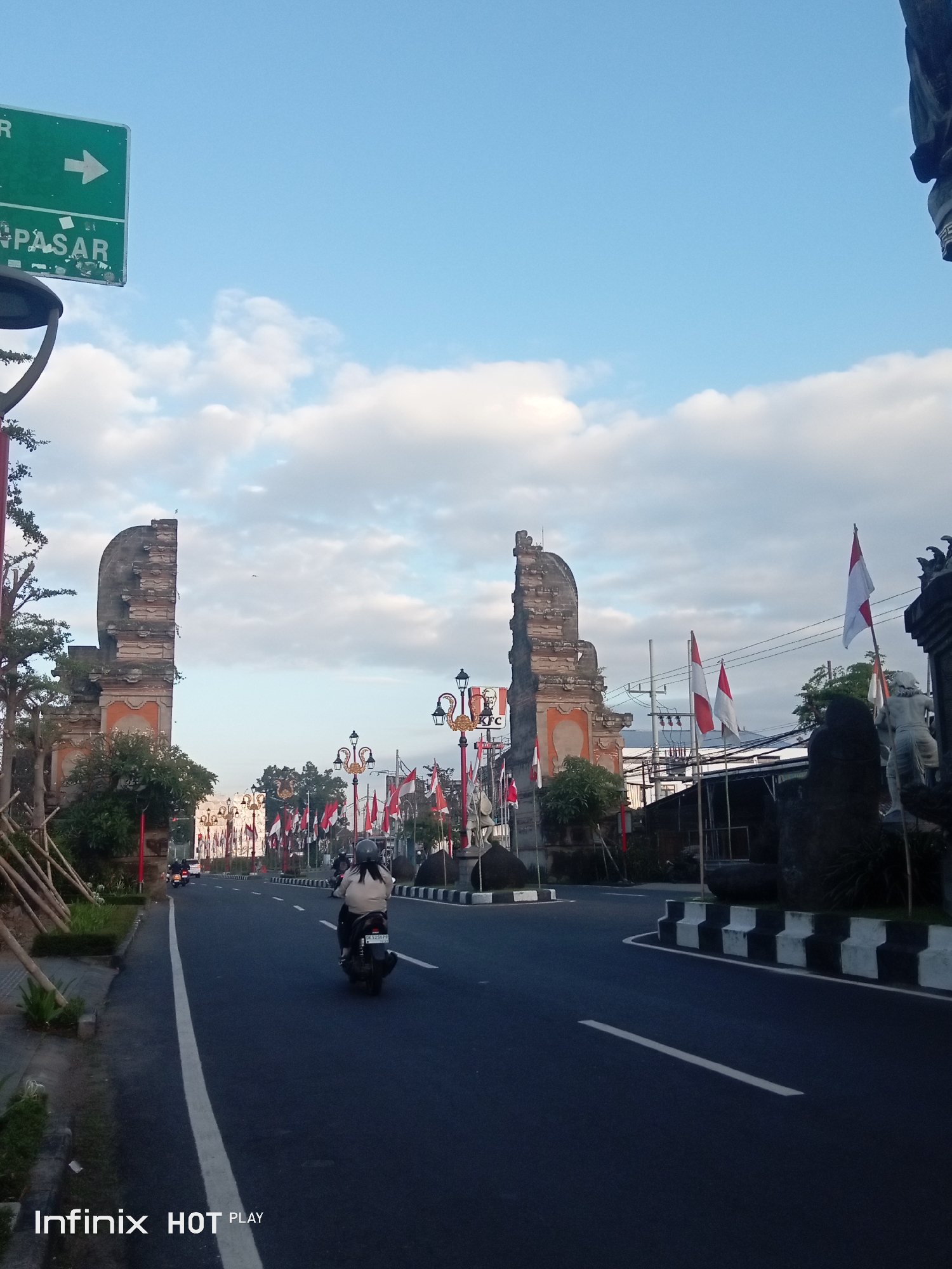 The image shows a street scene in Blahbatuh, Indonesia, with ornate archways on either side of the road. These archways appear to be part of traditional Balinese architecture, possibly marking an entrance or significant landmark. The road is paved and has a white lane divider. A single motorcycle with a rider is visible in the right lane, moving away from the camera. The rider is wearing a helmet and a light-colored jacket.
Along the sides of the road, there are decorative streetlights with ornate lanterns. Numerous Indonesian flags, red and white, are prominently displayed, suggesting a national celebration or observance. In the background, more of the traditional architecture and vegetation can be seen. The sky is partly cloudy with a bright, diffused light, indicating it might be early morning or late afternoon. A green directional sign with an arrow pointing to the right and the word "PASAR" is visible in the upper left quadrant of the image. The overall atmosphere appears serene and festive.