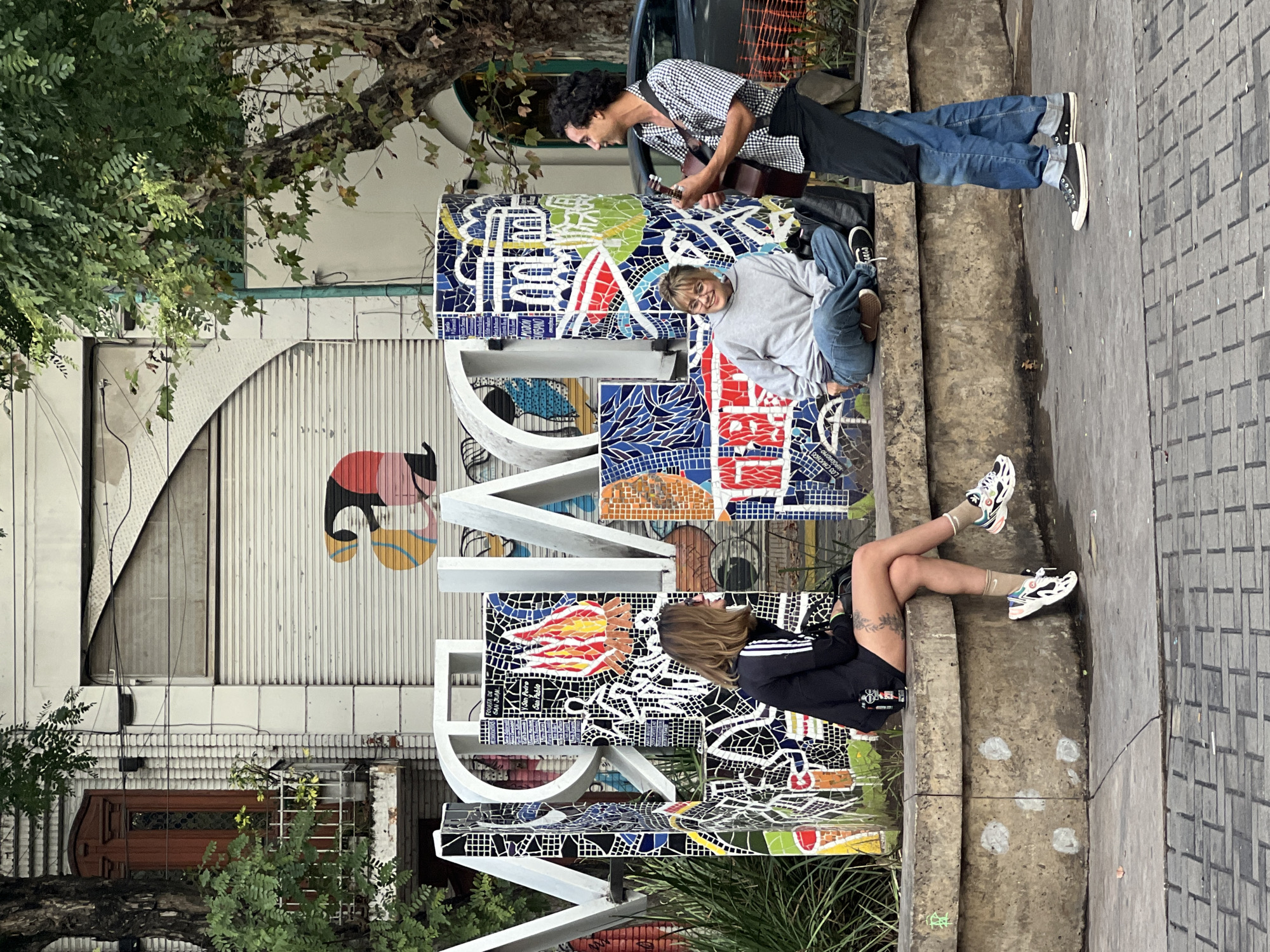 The image shows three young adults sitting on a tiled sign in Buenos Aires, Argentina. One person is playing a guitar, and the other two are seated next to them. The location is a public space in Buenos Aires, as indicated by the sign and the urban environment. The sign is large, three-dimensional, and decorated with colorful tiles. The building behind the sign has a rolling metal door.