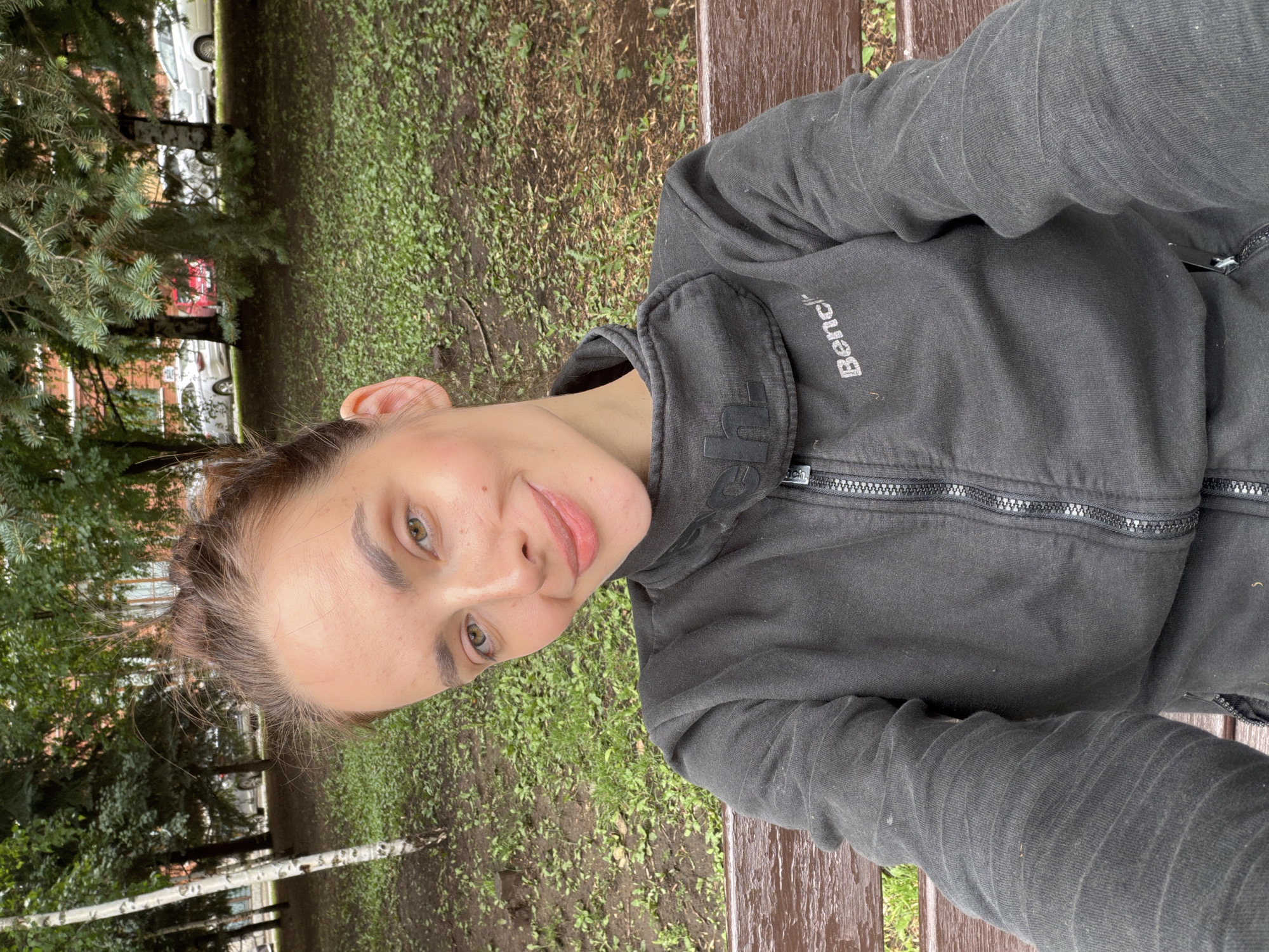The image is a close-up, selfie-style shot of a young woman with brown hair styled in a bun. She has light skin, light brown eyes, and a slight smile. She is wearing a dark grey zippered jacket with the brand "Bench" visible on the left chest. The jacket has a high collar. The woman is sitting on a wooden bench.
The background suggests an outdoor, urban environment. Behind the woman, there is a grassy area with scattered leaves and dirt patches. Several trees are visible, including a birch tree with white bark to the left and evergreen trees further back. In the distance, there are buildings with red brick facades and parked cars, indicating a residential or park area within a city. The overall lighting suggests an overcast day. The specific location within Ufa, Russia cannot be determined from the image alone.