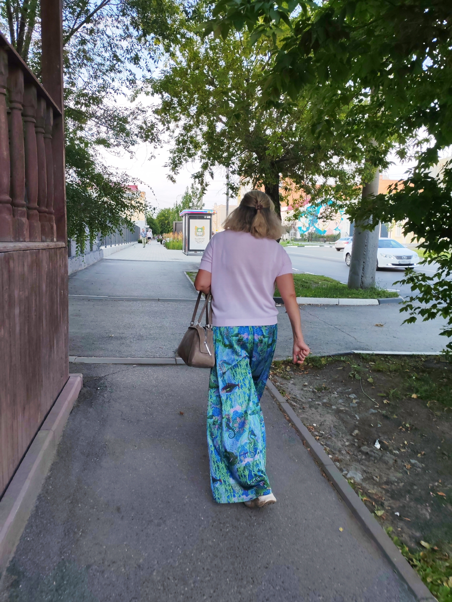 A woman with blonde hair pulled back is walking away from the camera on a paved sidewalk. She is wearing a light pink t-shirt and a long, flowing skirt with a blue, green, and white paisley pattern. She is carrying a light brown handbag over her left shoulder. To her left is a wooden fence and to her right is a grassy area and a street with cars driving. A bus stop shelter is visible further down the sidewalk. The scene appears to be in an urban environment with trees and buildings in the background.
