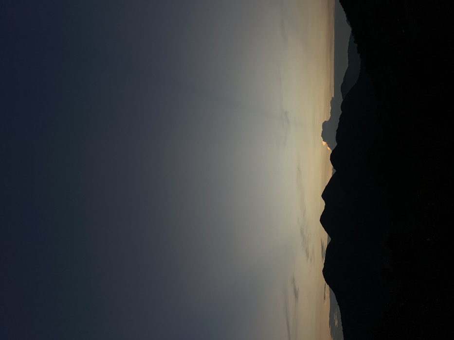 The image captures a mountainous landscape at twilight (dawn or dusk) in Nha Trang, Vietnam. The sky exhibits a vertical gradient, transitioning from dark grey-blue at the top to lighter hues of yellow and orange above the horizon. A prominent range of dark, silhouetted mountains dominates the lower half of the frame, their undulating peaks defining the horizon line. Behind these mountains, a single, illuminated cloud appears near the center-right, from which faint crepuscular rays are visible extending upwards into the lighter sky. No human subjects, man-made objects, or specific actions are present in the scene.