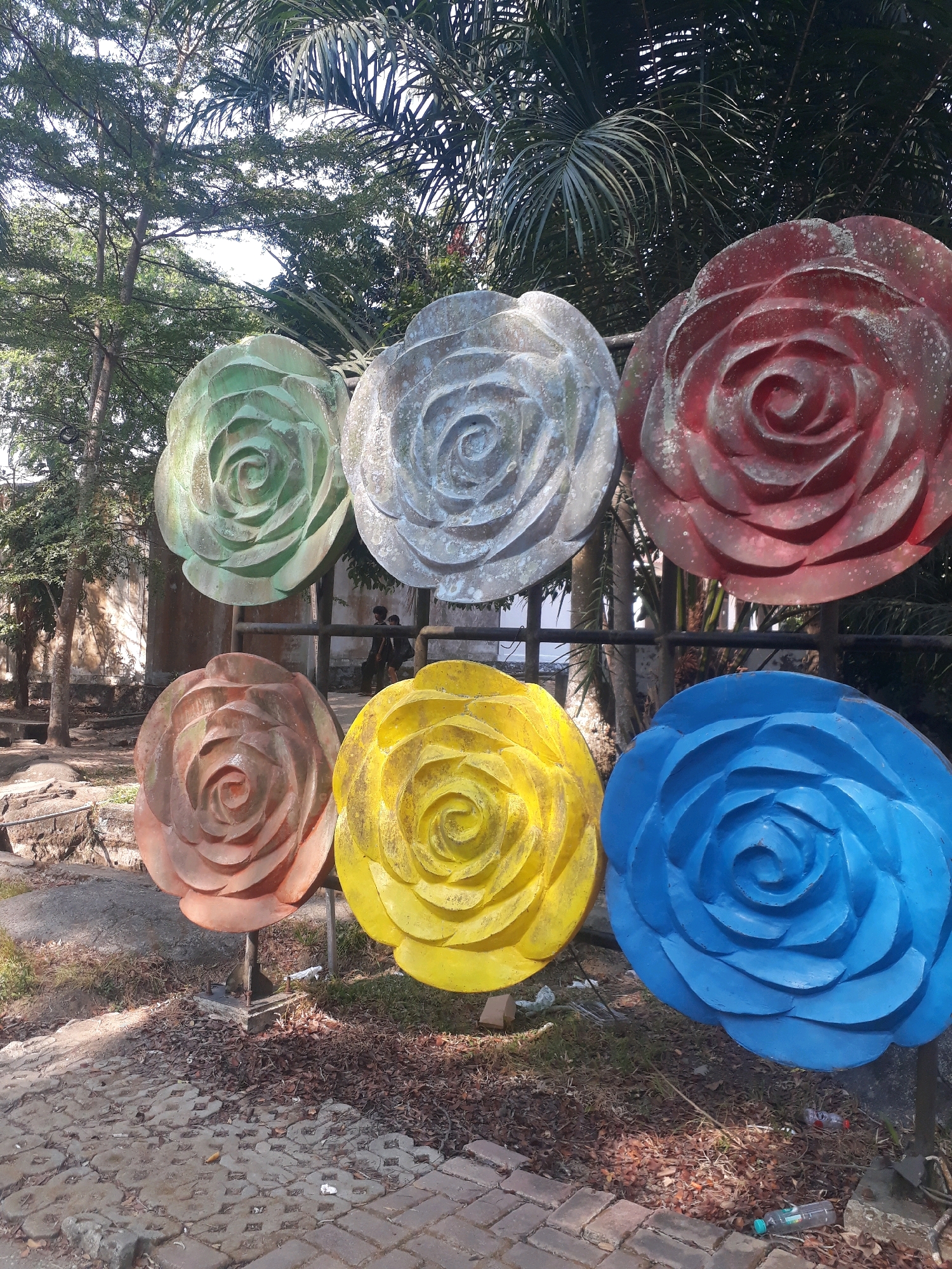 The image shows six large, colorful rose sculptures arranged in two rows of three. The top row consists of a green rose on the left, a light gray rose in the center, and a red rose on the right. The bottom row features a brown rose on the left, a yellow rose in the center, and a blue rose on the right. The sculptures appear to be made of concrete or a similar material and are weathered, with some dirt and discoloration visible.
Behind the sculptures, there is a black metal fence and a backdrop of trees and foliage, including palm fronds. Sunlight filters through the leaves, creating dappled shadows on the ground. In the middle ground, between the rows of roses, two individuals are visible, blurred due to their movement and the focus of the image. They are walking past the sculptures, one appearing to be wearing a dark backpack.
The foreground of the image shows a paved area made of interlocking concrete bricks, with some areas covered in dry leaves and sparse grass. The overall setting suggests an outdoor public space, possibly a park or a decorative area within a facility.
This scene is located at the Taman Sari Water Castle in Pangkalpinang, Indonesia. This historic site, also known as Taman Sari Pangkalpinang, features remnants of a former royal bathing complex and gardens. The rose sculptures are a modern addition, likely intended to enhance the visual appeal of the area.