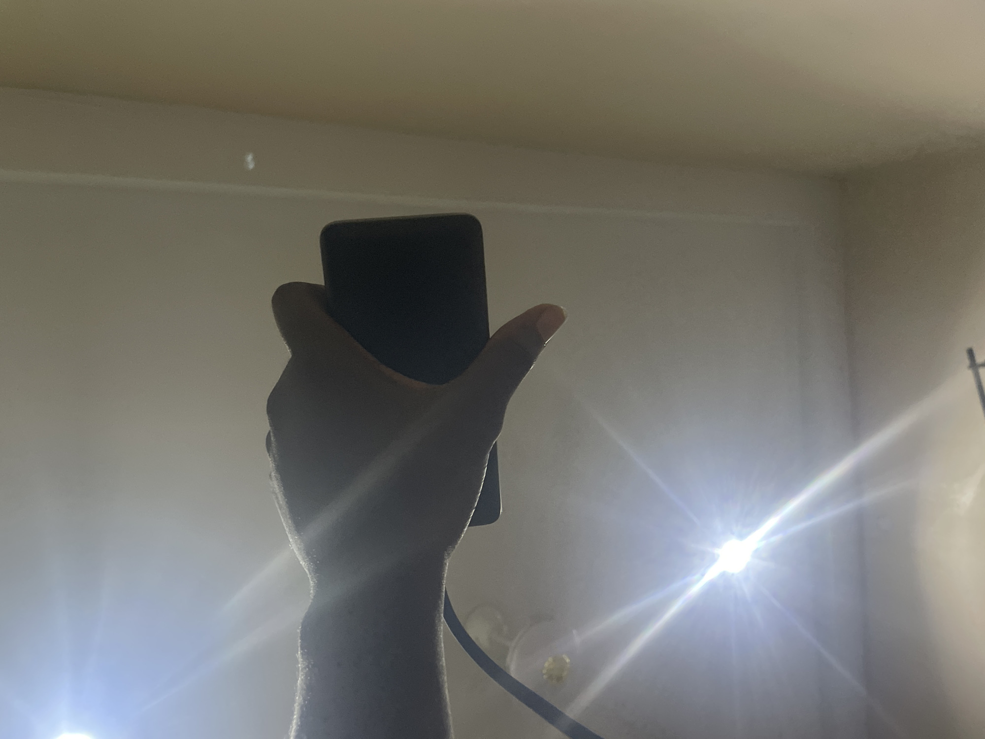 A close-up, low-angle shot shows a person's hand holding a dark, rectangular object, likely a smartphone, horizontally. The hand is dark-skinned, with the thumb on the bottom and fingers curved around the object. A bright light source, possibly a flash or overhead lamp, is positioned in the upper left quadrant of the image, creating a lens flare and washing out parts of the background with intense light. The background appears to be an indoor environment with walls that are dimly lit and off-white or beige in color. The scene is very dark overall, with significant glare from the light source. This image was captured in Jalingo, Nigeria.