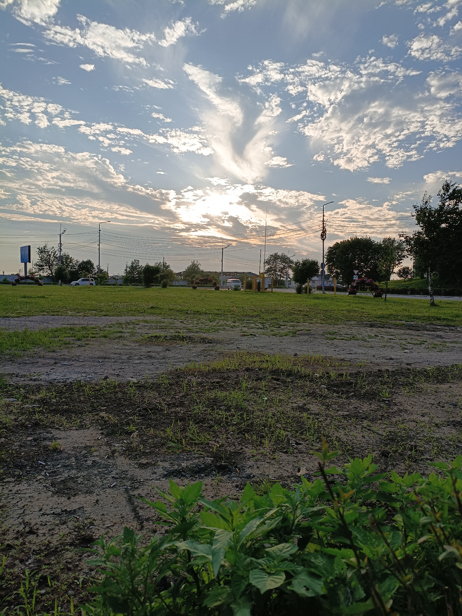 The image captures a wide-angle view of a grassy area with patches of dirt in the foreground, leading to a paved road in the mid-ground. The sky above is filled with dramatic clouds, with the sun peeking through, casting a warm light. Along the road, there are trees, streetlights, and a few cars, suggesting an urban or suburban setting. The overall impression is of a peaceful, possibly early morning or late afternoon scene. The location is identified as Korsakov, Russia, which provides a geographical context for the image.