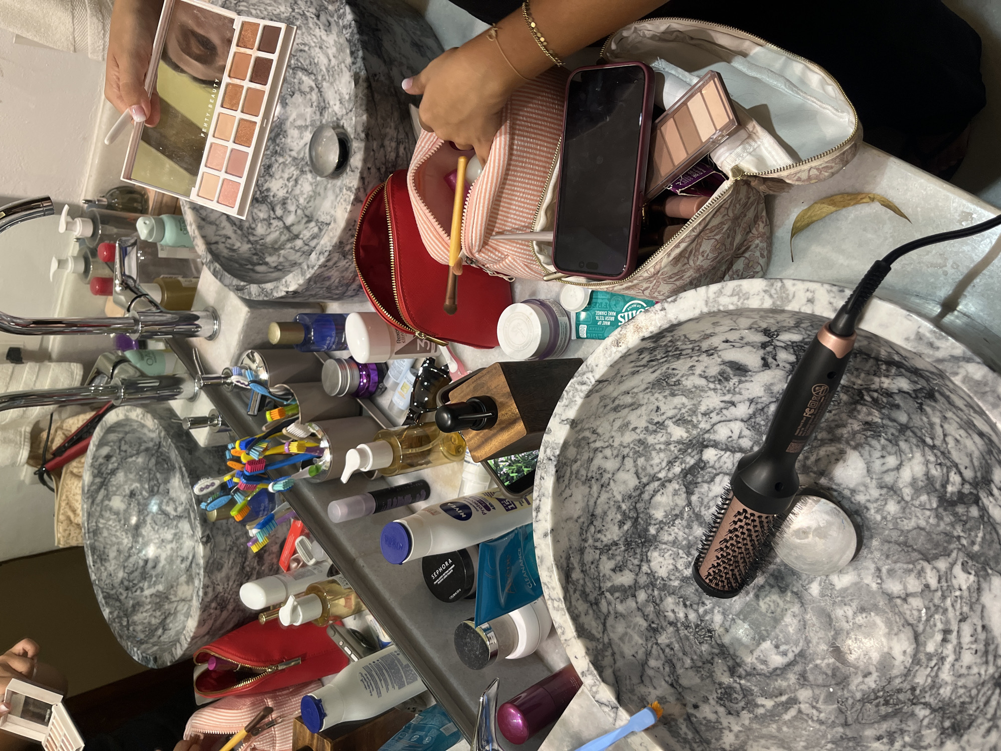 The image depicts a bathroom vanity with a collection of personal care and beauty products arranged around two marble sinks. A person's hands are visible, holding an open makeup palette, specifically a Fenty Beauty eyeshadow palette with shades of beige, tan, and brown. The palette is positioned over a sink.
On the vanity, several cosmetic items are present, including makeup brushes, various bottles of skincare and haircare products, toothbrushes in a cup, and a hairdryer. A smartphone is also visible, placed on top of a cosmetic bag. The environment appears to be a well-lit bathroom, possibly in a hotel or residential setting, given the marble countertops and sinks. The overall impression is one of a personal grooming or makeup application session in progress. There are no specific indicators of the city of Huixquilucan de Degollado, Mexico, within the image itself.