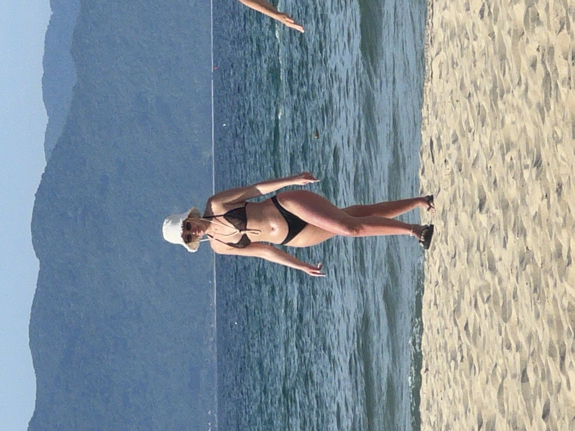 In Đà Nẵng, Vietnam, on a sandy beach adjacent to the ocean, a woman wearing a white bucket hat and a dark bikini walks towards the viewer. The ocean is a vibrant blue, with gentle waves lapping the shore. In the background, lush green mountains rise against a clear sky. To the right of the frame, another person's arm and hand are visible, extending towards the ocean. The scene captures a serene moment of leisure by the sea.