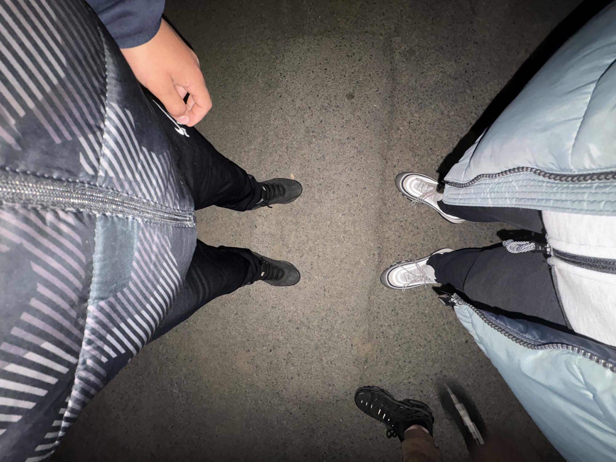 The image depicts a low-angle, overhead view of three people standing on a paved surface. The person on the left wears a dark jacket with a geometric white and gray pattern and black pants and gray shoes. The person on the right wears a light blue puffer jacket, black pants, and silver shoes. The third person, at the bottom, wears black shoes and light brown pants. The setting appears to be outdoors at night. The location, Goritsa, Bulgaria, cannot be confirmed based on the image alone.