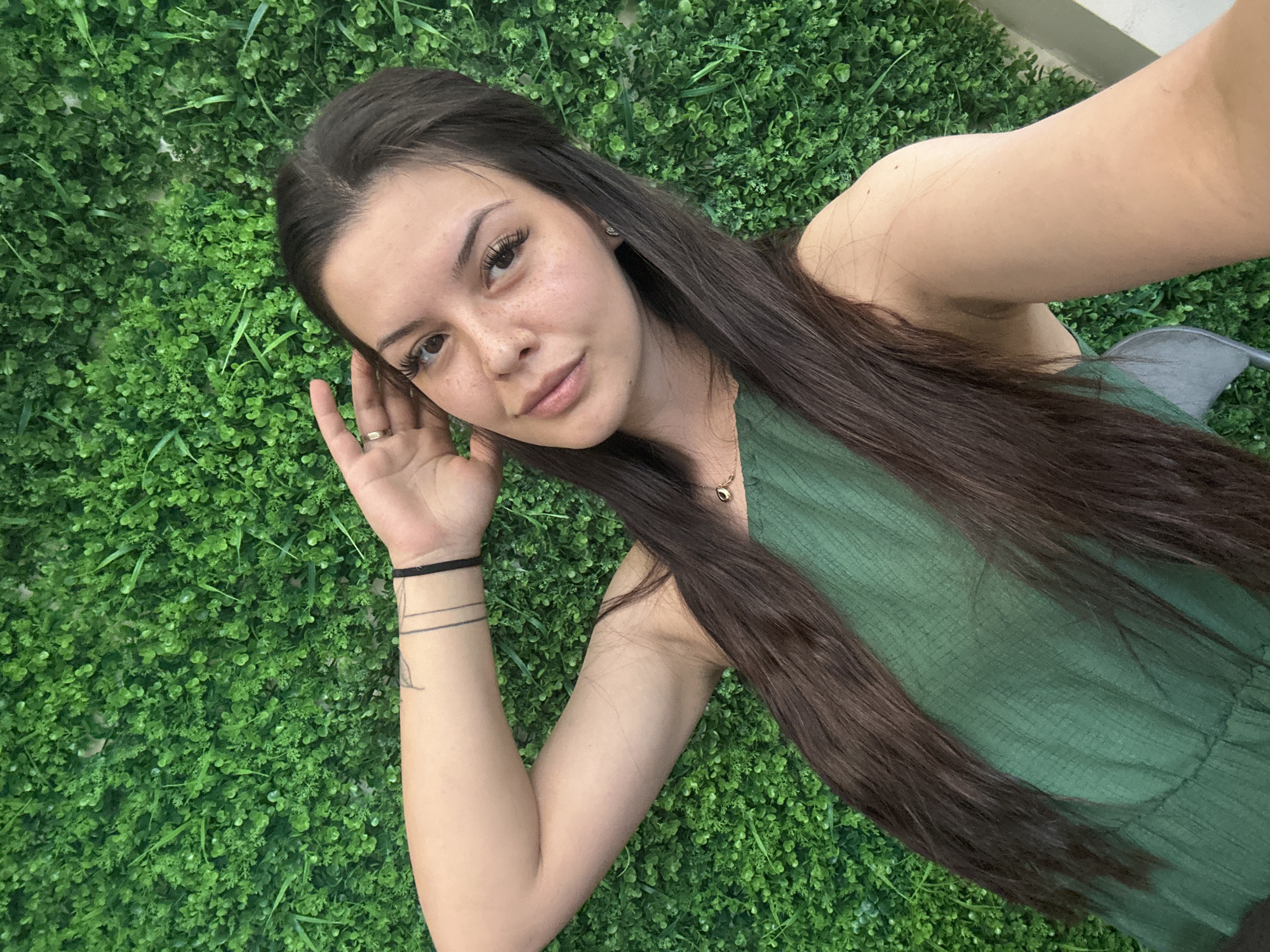 A young woman with long, dark hair and light skin is lying down in front of a green, artificial grass wall. She is wearing a green, sleeveless dress and a gold necklace. Her right arm is raised and bent, with her hand resting on her head. She is looking at the camera with a slight smile. Her left arm is bent at the elbow and her hand is resting on her chest. She has a few freckles on her nose and cheeks and is wearing a ring on her right hand. She also has a black bracelet and a tattoo on her left wrist. The setting is an indoor or outdoor space with a prominent, textured green wall, likely in Sliven, Bulgaria, given the context provided.