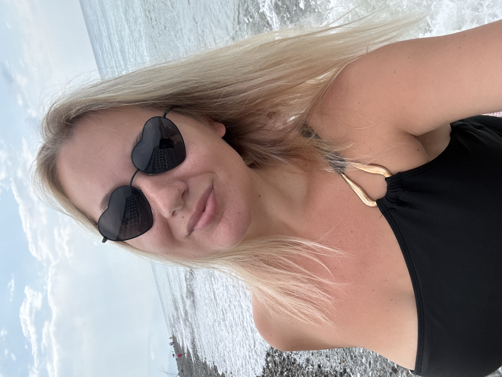A blonde woman in a black swimsuit and heart-shaped sunglasses takes a selfie on a rocky beach with the sea behind her. The sea is choppy with white foam. In the background, people are visible on the beach and in the water. The sky is cloudy. The image appears to have been taken in Giachrypsh, Abkhazia, based on the context provided.