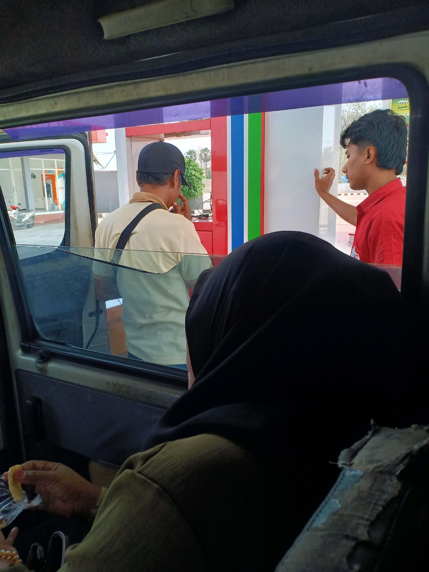 In Semanding, Indonesia, at a gas station, a person in a red shirt appears to be interacting with a customer in a beige shirt who is standing outside a vehicle. The customer, wearing a black baseball cap, is facing away from the camera. The gas pump has vertical stripes of red, blue, green, and white. Inside the vehicle, a person wearing a black hijab is visible, and another person's hand is holding a piece of food. The overall scene suggests a typical transaction at a gas station, with elements of everyday life captured within the frame.