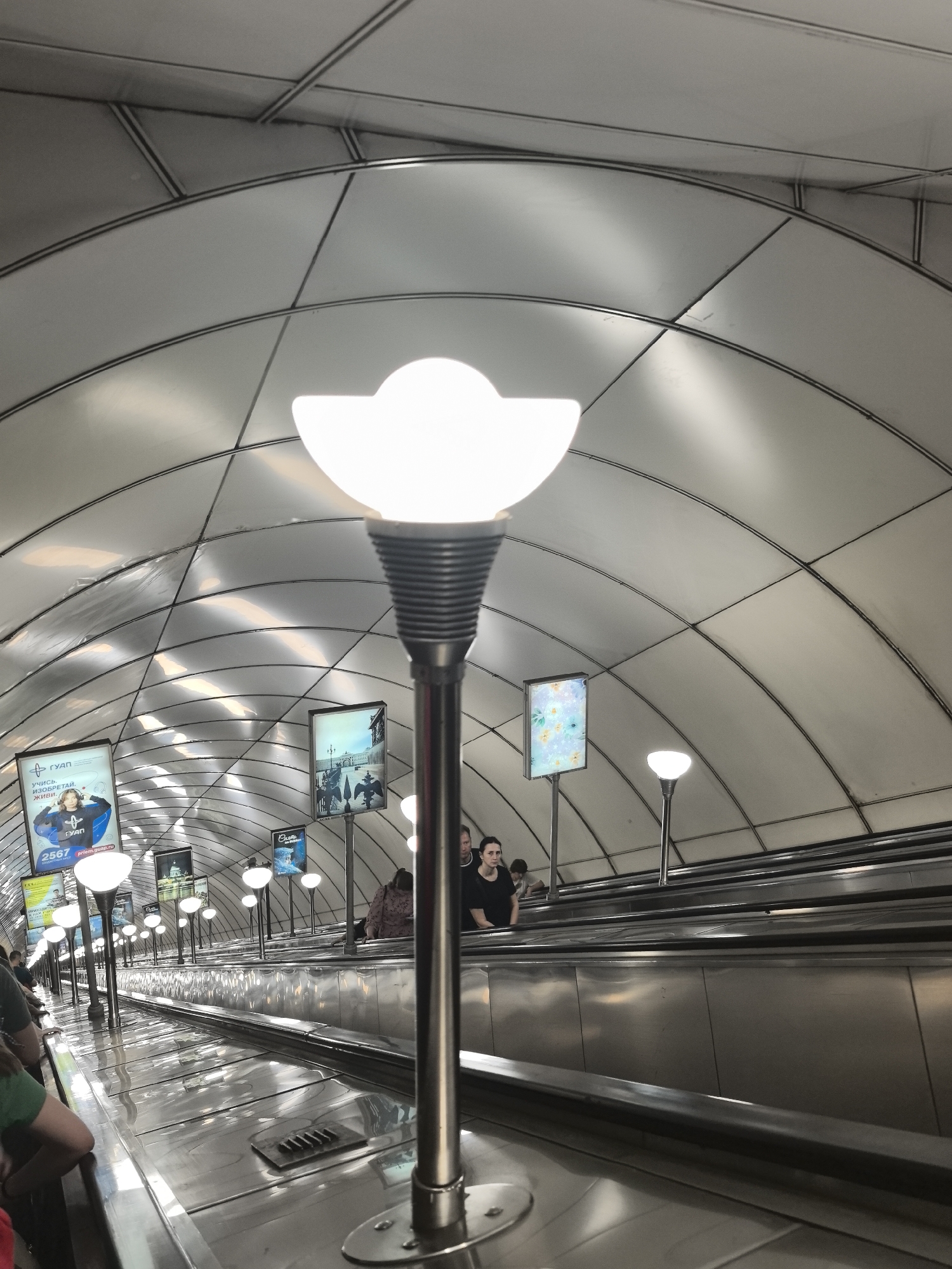 In Saint Petersburg, Russia, this image captures an upward-facing escalator in a metro station. The ceiling is a striking feature, composed of curved, metallic panels arranged in a repeating pattern, with visible structural beams. Along the escalator's sides, illuminated advertising displays are mounted on poles, some featuring text in Cyrillic script. Several people are present, some on the escalator and others standing near the top. The escalator itself is made of polished metal, reflecting the ambient light. A prominent light fixture, with a distinctive white, petal-shaped shade, stands in the foreground, its metallic pole extending upwards. The overall environment is modern and functional, characteristic of a well-lit urban transportation hub.