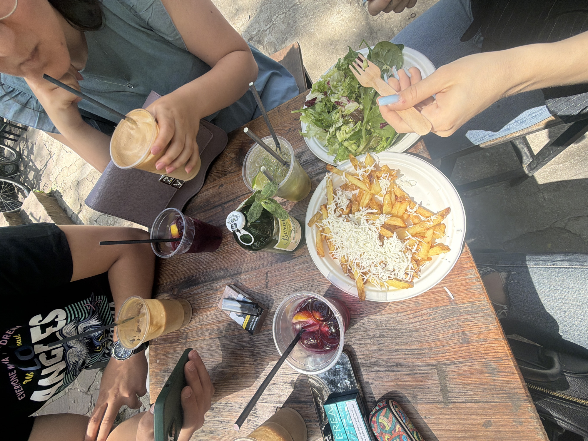 The image shows a group of people seated around a weathered, wooden table on what appears to be a sunny day. The scene is set at an outdoor dining area, possibly in a cafe or restaurant. The location in Sofia, Bulgaria cannot be verified from the given image.
Several individuals are visible with portions of their bodies present, including hands, arms, and parts of their torsos. One person is wearing a light blue top and another wears a black t-shirt with graphic print.
The table is covered with an assortment of food and drinks, including a plate of fries topped with shredded cheese, a salad, and several clear plastic cups filled with beverages. The drinks range from iced coffees and a dark red drink to a mojito. A bottle of what appears to be a soft drink is also visible. Additionally, there are mobile phones, a pack of cigarettes, and a small colorful patterned case on the table.
One individual is holding a wooden fork over the salad. Another appears to be interacting with a mobile phone. The presence of straws in the drinks suggests the act of consuming these beverages. The overall action taking place appears to be a social gathering for food and drinks.
The environment shows dappled sunlight cast on the table and surrounding areas. The quality of the light and shadows suggests that it's a sunny day. The setting implies an urban environment, given the outdoor dining setting.
