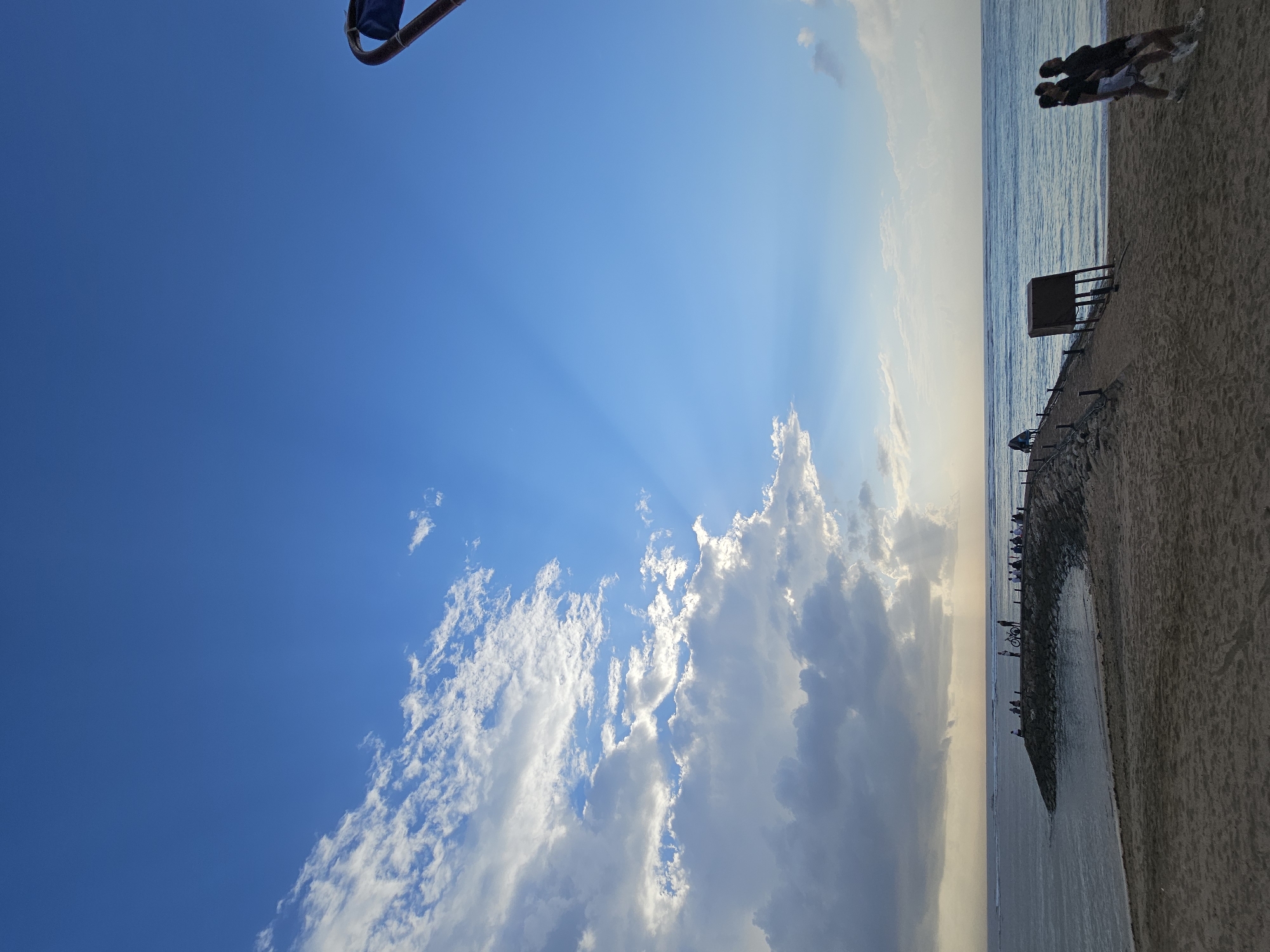 The image depicts a coastal scene at sunset in Denpasar, Indonesia. A sandy beach in the foreground transitions to a rocky breakwater extending into the ocean. Several people are visible on the breakwater, some standing and others sitting, appearing to observe the sea or engage in conversation. A bicycle is parked on the breakwater. In the middle ground, the ocean stretches to the horizon under a vast, partly cloudy blue sky. Sunbeams are piercing through a gap in the clouds, creating a dramatic effect. On the far right of the image, a dark, angular structure resembling a shelter or signpost stands on the beach. In the foreground on the beach, three individuals are walking away from the camera. The overall atmosphere is serene and suggests a popular spot for locals and tourists to enjoy the evening.