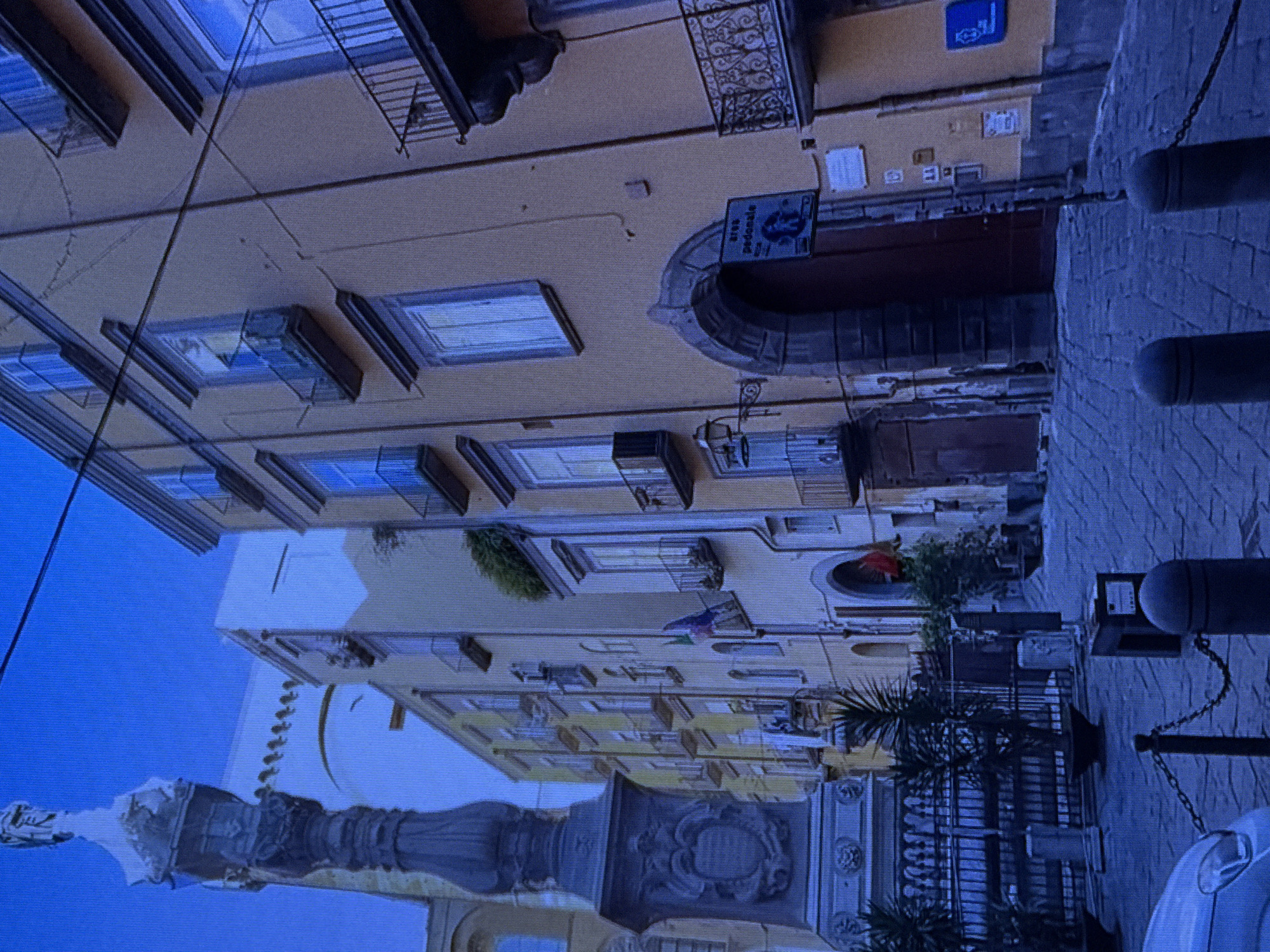 The image depicts a European city street with narrow cobblestone paving and tall, narrow buildings painted in warm, muted colors such as yellow and cream. A dark, intricately carved monument stands prominently on the left side of the frame. The buildings feature numerous windows with dark balconies and shutters, some of which are closed. Wires are visible stretched across the sky above the buildings.
On the right side of the street, an arched doorway is visible, above which hangs a sign indicating a pedestrian area ("area pedonale"). There are also several flags, one of which appears to be the Italian flag, hanging from the buildings. A metal fence with planters containing greenery is present in the foreground, and a car is partially visible on the lower left. The scene is devoid of people and appears to be captured during daylight hours, though the sky is a deep blue suggesting it may be late afternoon or early morning. The architectural style suggests a historical European city, possibly in Italy.