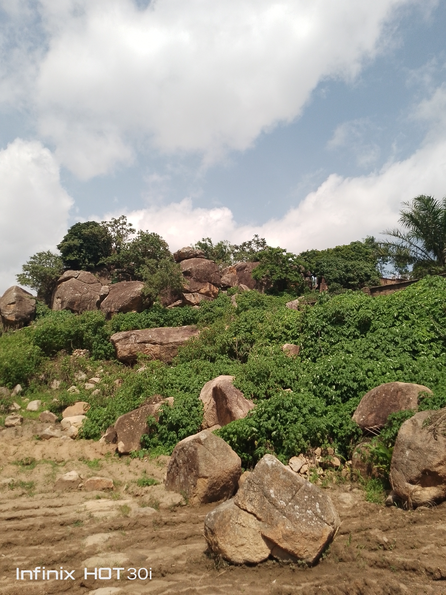 A rocky, vegetated hillside under a cloudy sky. The foreground consists of a dirt path with scattered rocks and some tire tracks. The midground is a steep incline covered with lush green vegetation and large, weathered rocks. In the background, more rocks and trees are visible, with a palm tree on the far right. The sky is blue with scattered white clouds. The bottom left corner of the image contains a watermark that reads "Infinix HOT 30i". There are no people or animals visible in the image.