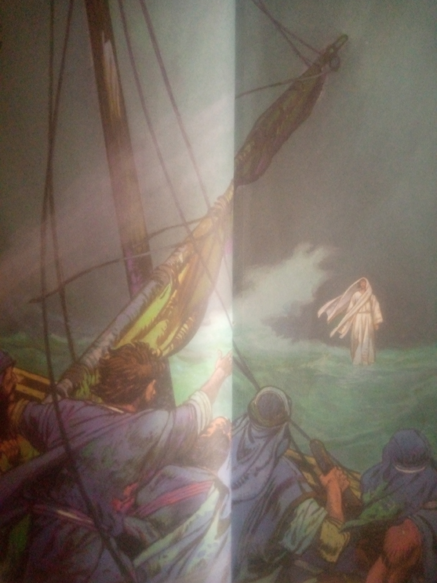 This is an illustration depicting a dramatic scene at sea. The primary subjects are a group of people on a boat and a lone figure walking on the water. The boat appears to be a sail boat, with a prominent mast and rigging visible. Several individuals are shown on the boat, some appearing to be in distress or awe, with their arms raised or expressions of surprise. Their clothing suggests a historical or biblical context, with draped garments.
The lone figure, standing on the turbulent water, is depicted in a white robe, also with a draped appearance, and appears to be walking towards the boat. The sea is rough, with visible waves and spray, suggesting a storm or rough weather. The overall mood of the illustration is one of wonder, fear, and perhaps relief, given the presence of the figure on the water.
The lighting is dramatic, with a sense of mist or fog obscuring parts of the scene, particularly towards the left and background, creating a somewhat ethereal atmosphere. The colors are muted in some areas due to the mist, while the figures and the water are rendered with more vibrancy.
There is no visible text in the image. The scene evokes a biblical narrative, likely the account of Jesus walking on water, given the visual elements. The location context of Jauro Yinu, Nigeria, is not visually represented in the illustration itself and appears to be external information provided for reference.