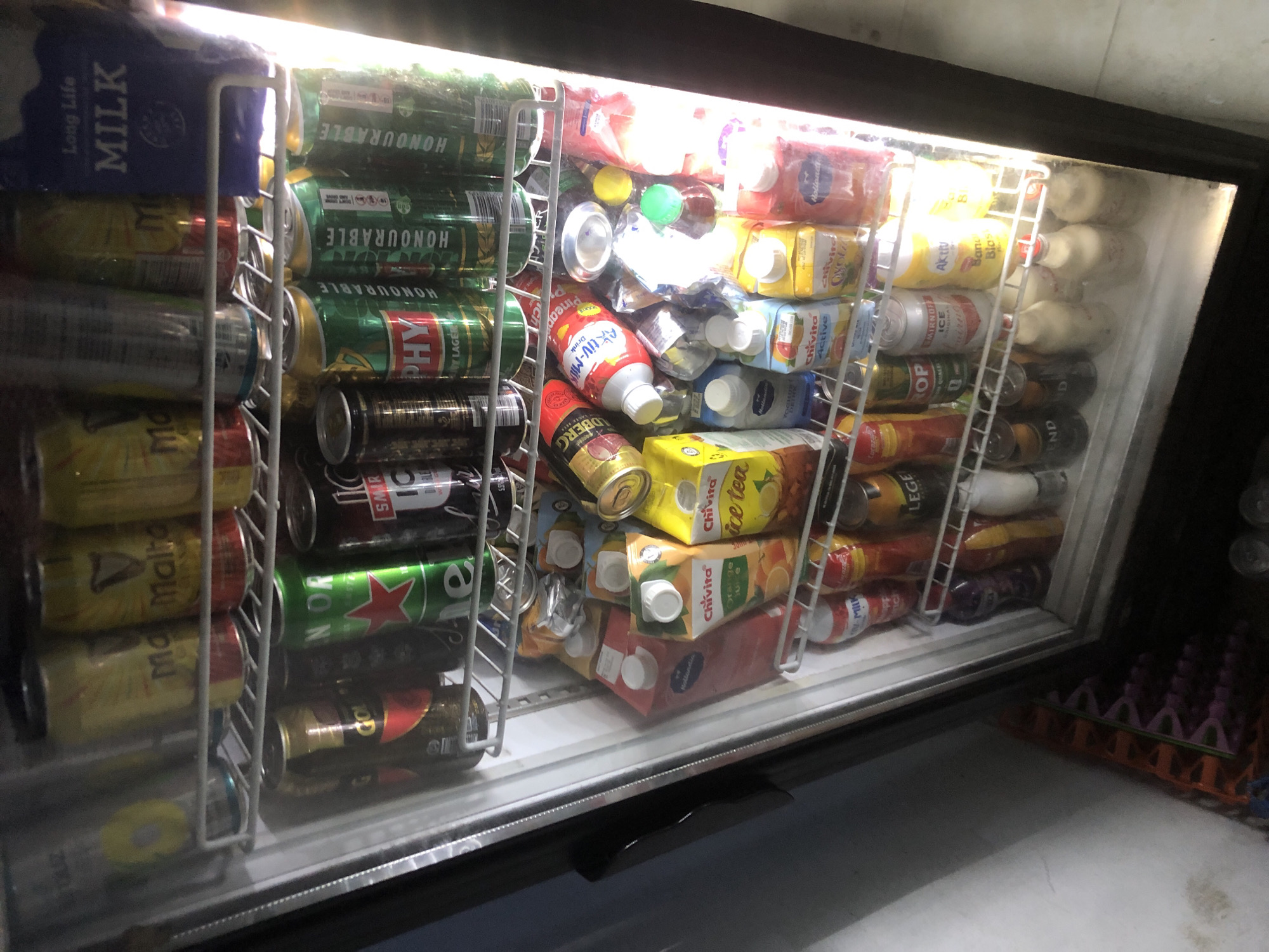 The image shows a commercial refrigerator stocked with beverages, primarily in cans and cartons. The refrigerator is positioned indoors, with a bright light emanating from within, illuminating the contents. The shelves are organized with rows of drinks. On the top shelf, there are several yellow and gold colored cans, some with visible labels such as "Malta Guinness". To the right, a blue carton with the text "Long Life MILK" is visible. The second shelf contains green cans with red stars, likely depicting a brand of beer or soft drink. There are also some darker colored cans and what appears to be a bottle of Smirnoff Ice. The third shelf is filled with various juice cartons, including "Chivita Ice Tea" and "Chivita Orange Juice", as well as some red cartons. Below these, on the fourth shelf, are more cans and bottles, including what looks like a bottle labeled "Active Milk Drink" and some bottles of what appear to be alcoholic beverages. The bottom shelves show more bottles of milk or dairy drinks, and some darker colored bottles. The overall impression is a retail display of cold drinks in a store or supermarket. The context of Ado, Nigeria, suggests this is a point of sale for beverages in that region.