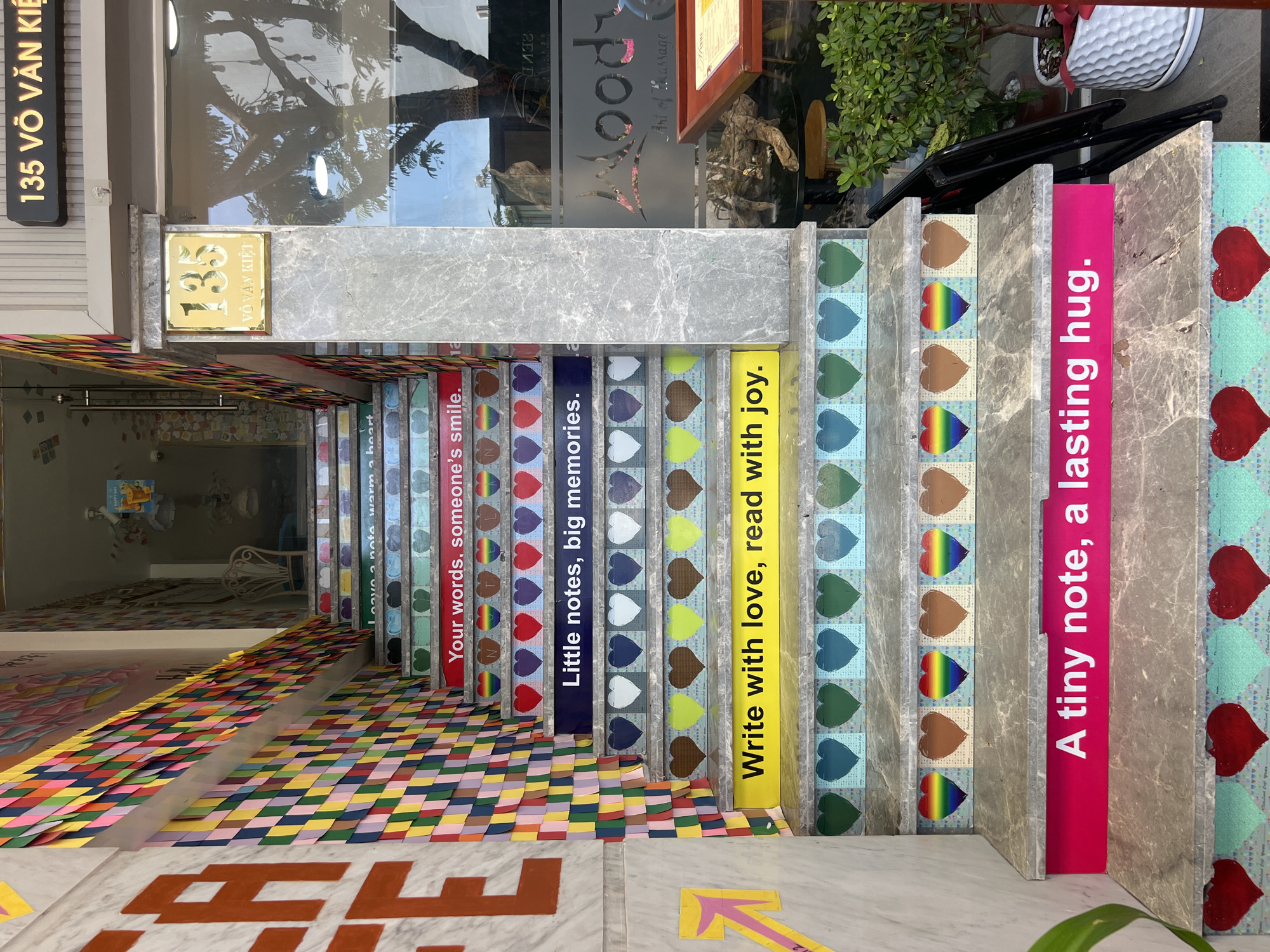 The image shows a set of outdoor stairs decorated with colorful hearts and positive messages. The stairs are made of grey marble and each step is decorated with a repeating pattern of hearts in various colors and shades. Some steps have colorful rainbow hearts, while others have solid colored hearts in shades of blue, green, brown, and red.
The risers of the stairs are painted with different colored backgrounds, and each step has a phrase written on it in white or black lettering. From bottom to top, the visible phrases are: "A tiny note, a lasting hug.", "Write with love, read with joy.", "Little notes, big memories.", "Your words, someone's smile.", and "Leave a note, warm a heart."
The left side of the stairs is adorned with a wall covered in colorful strips of paper arranged in a diamond pattern. To the right of the stairs, there is a glass window with the text "oody Art of Massage" visible. A sign above the window reads "135 VÕ VĂN KIỆT".
The overall impression is a vibrant and welcoming entrance, likely to a business or establishment. There are no people visible in the image.