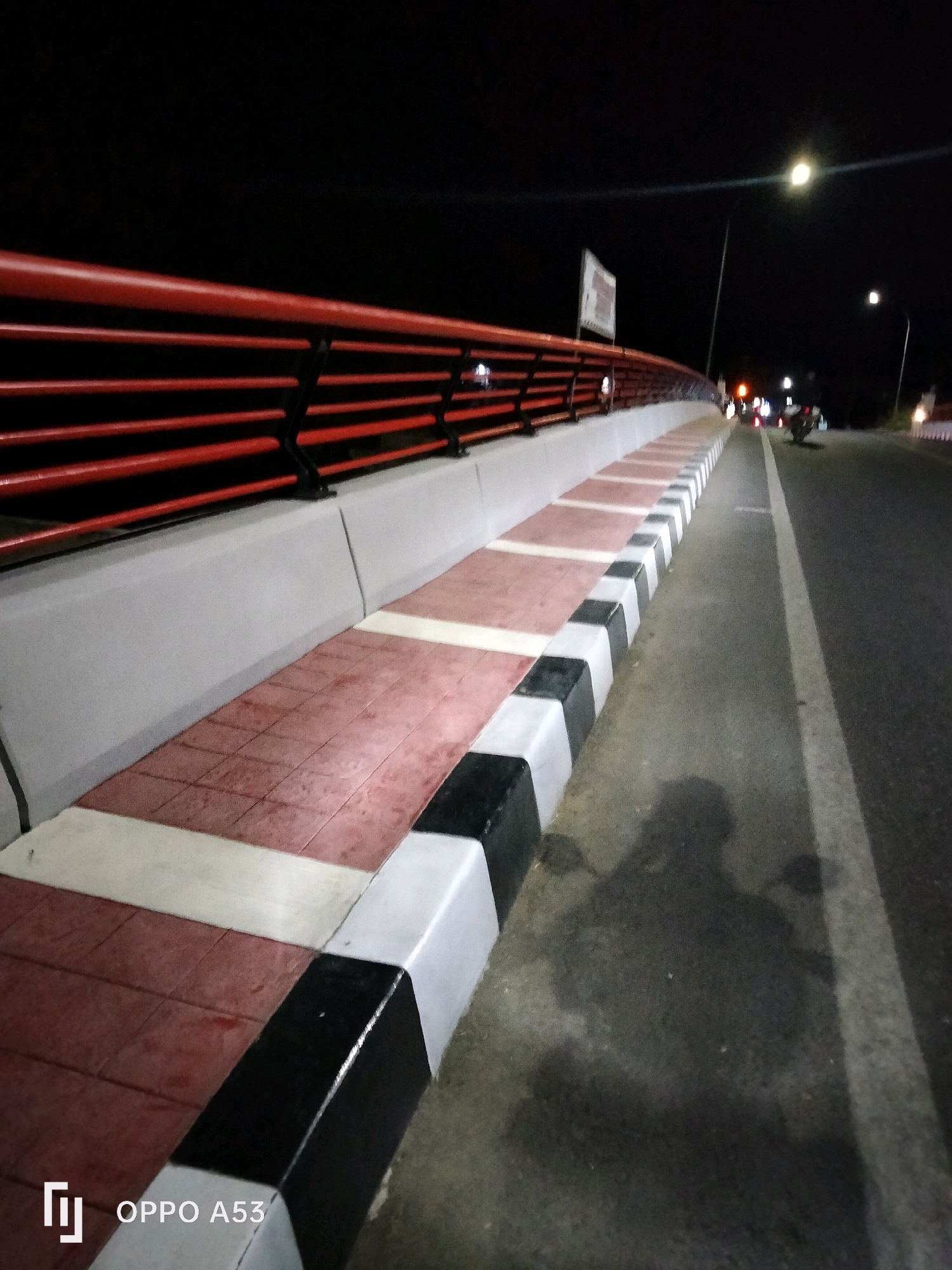 The image depicts a street scene at night in Bendo, Indonesia, with a focus on a pedestrian walkway alongside a road. The walkway features a distinct pattern of red, white, and black tiles, bordered by white concrete curbs that are painted with black accents. A red railing with horizontal bars runs along the pedestrian side of the walkway. The road itself is paved and has a white line marking its edge. Streetlights illuminate the scene, casting light on the road and walkway. In the background, there are indications of vehicular traffic with visible headlights and taillights. A shadow of a person, likely riding a motorcycle, is cast on the walkway. The overall environment is dark, characteristic of nighttime, with artificial lighting providing visibility.