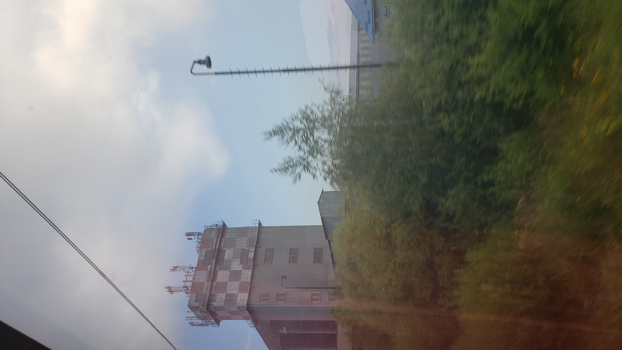 The image captures a view from inside a vehicle, likely a train, looking out at an industrial area. The dominant structure is a tall, checkered tower, possibly an old control tower or industrial building, with a predominantly grey facade and alternating red and grey squares on its upper section. Several antennas are visible on its roof. To the right of the tower, a tall, slender lamppost with a curved head stands against a pale sky with scattered clouds. Beyond the tower, a long, low-lying industrial building with a grey roof stretches into the distance, and to its right, another building with a blue roof is partially visible. A dense line of green foliage, including trees and bushes, runs across the foreground, creating a blurred effect due to the motion of the vehicle. The overall scene suggests an industrial or commercial district on the outskirts of a city, consistent with a location in Poprad, Slovakia, known for its industrial zones. The image is taken during daylight hours.