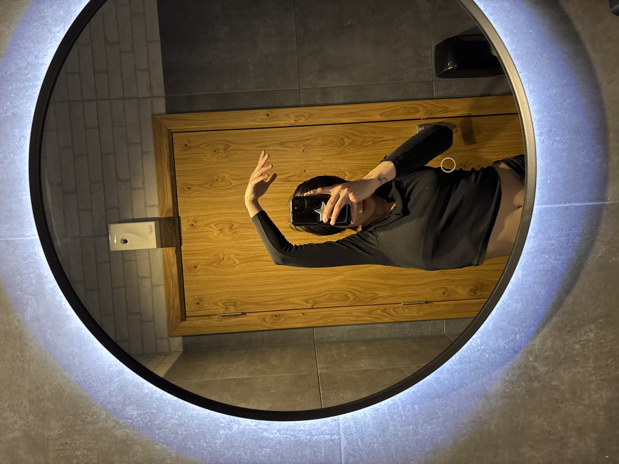 A person in black clothing is holding a phone with a star-shaped sticker in front of their face, taking a mirror selfie. The mirror is circular and has a lit-up, diffuse white light around the edge. The background reflected in the mirror includes a wooden cabinet or door, gray tiled flooring, and white brick walls. A soap dispenser is also visible on the brick wall. The location is unknown, but the description of the tiles, brick wall, soap dispenser, and mirror don't provide enough information to determine the city or country.