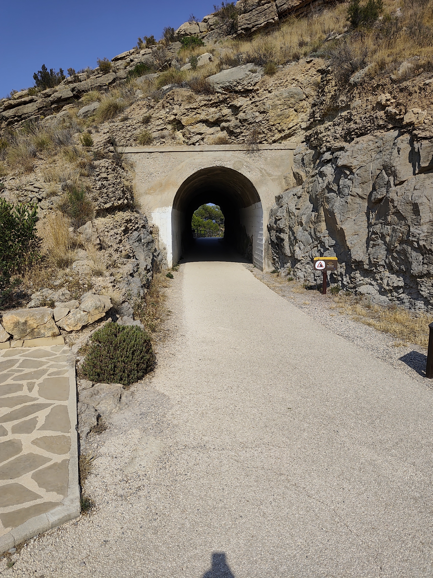 The image depicts a pathway leading towards a tunnel carved into a rocky hillside. The pathway is paved with light-colored gravel, and on the left, there is a section paved with irregular, flat stones. A small, bushy green plant grows near the edge of the stone paving. The tunnel entrance is arched and made of concrete, with visible stonework on the interior walls. Beyond the tunnel, the scene opens up to a sunlit area with trees and a railing. The hillside is covered in sparse vegetation and rocks, with a clear blue sky above. A brown sign with a yellow top and a circular warning symbol is visible on the right side of the path, near the tunnel entrance. The lighting suggests it is daytime, with a shadow of a person visible on the gravel path in the foreground.