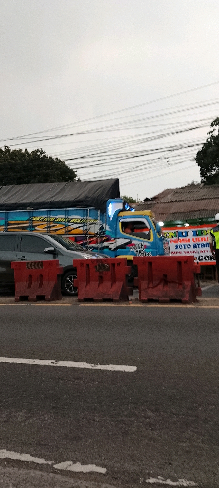 In Depok, Indonesia, on an overcast day, a blue and white truck adorned with colorful decals is parked on a road. The truck is equipped with flashing blue lights on its roof. In front of the truck, a gray car is stopped. Red plastic barriers are placed on the road, segmenting the scene. A white banner with Indonesian text, including "SOTO AYAM" and "NASI UDU," is visible behind the truck, suggesting a food vendor. The environment includes trees in the background and power lines crisscrossing the sky. The road has white lane markings.