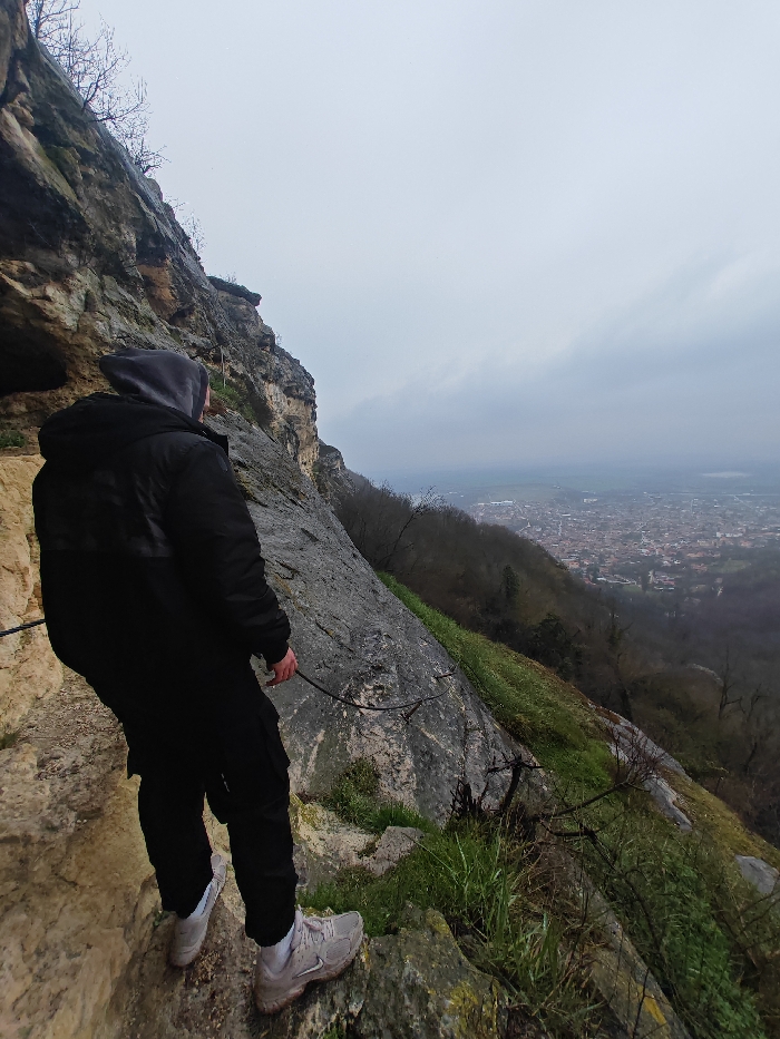 One individual, viewed from the back, stands on a rocky, elevated ledge overlooking the city of Shumen, Bulgaria. The person is wearing a black hooded jacket with the hood up, dark trousers, and light-colored athletic shoes. Their left hand is near a dark metal safety cable or rope secured along the adjacent steep, textured rock face, which transitions from light beige to dark grey. The right side of the frame shows a downward-sloping rocky terrain interspersed with green grass and sparse, bare-branched vegetation. In the background, the city of Shumen extends across a valley or plain, densely packed with buildings featuring reddish roofs. The sky is uniformly overcast and light grey, suggesting cool, cloudy weather conditions.