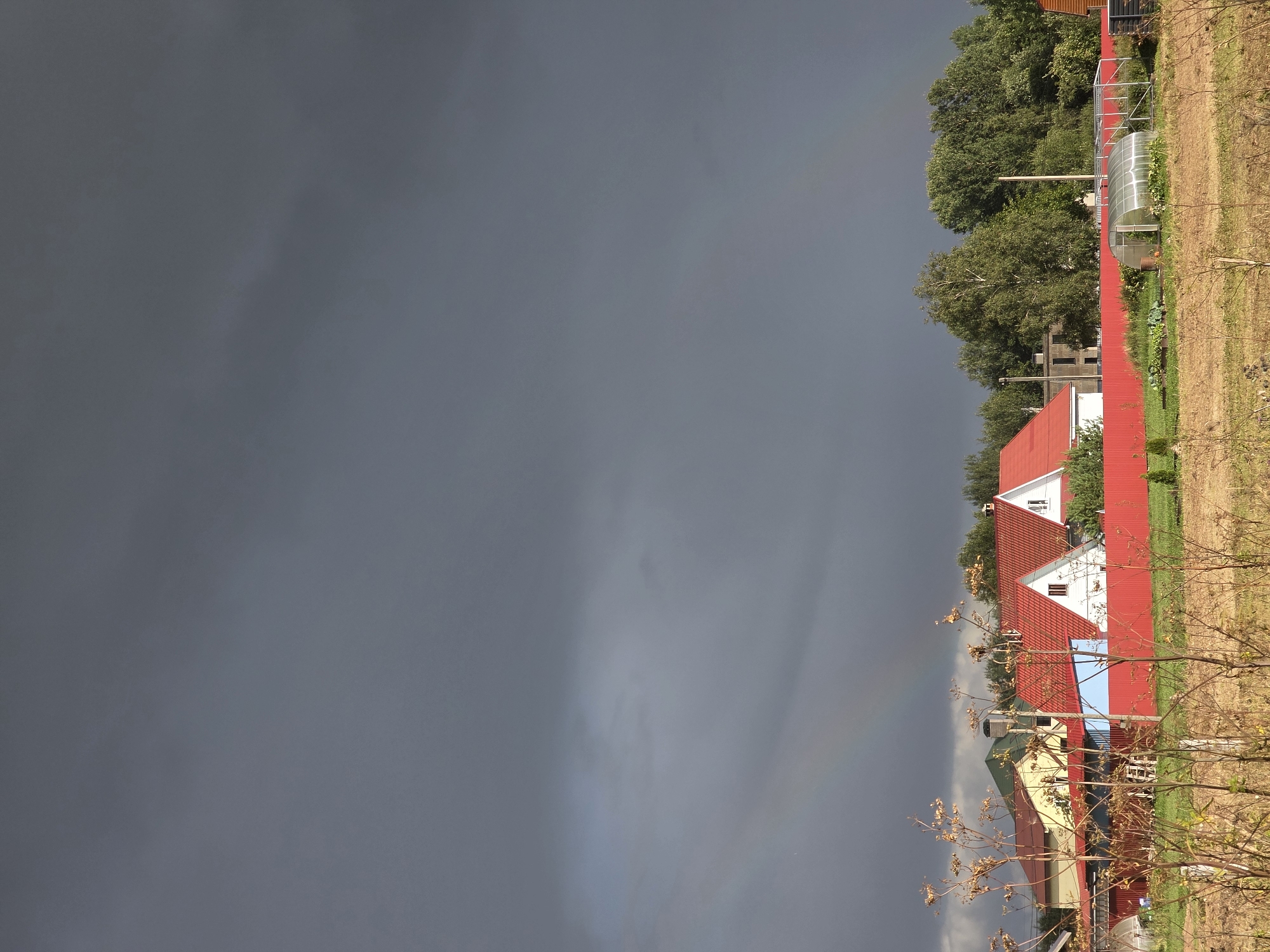 In the city of Glukhovo, Russia, a rural scene unfolds under a dramatic, dark gray sky. A faint rainbow arcs across the lower portion of the sky, hinting at recent or impending rain. In the mid-ground, a row of houses with red tiled roofs and white walls are visible, separated by a vibrant red metal fence. A bright blue section of a house adds a splash of color. To the right, a small greenhouse with a metal frame and translucent covering sits amongst green grass and garden beds. Behind the houses and greenhouse, a dense line of lush green trees forms a backdrop. In the foreground, dry, brown grass and thin, twig-like vegetation are in sharp focus, suggesting a dry field. The overall atmosphere is one of rustic tranquility juxtaposed with the imposing, stormy sky.