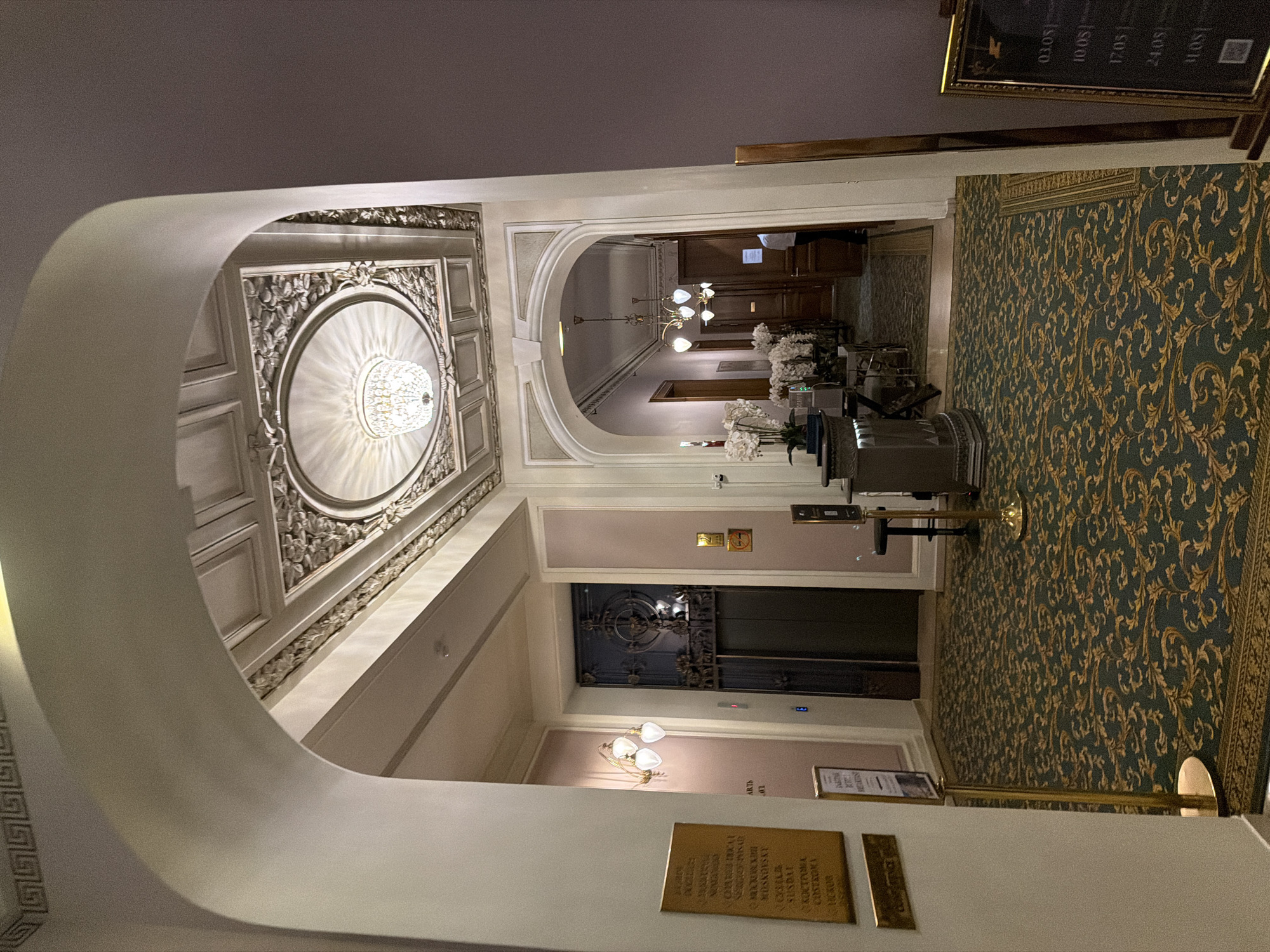 The image captures an interior view of what appears to be a high-end hotel or grand building in an unidentified city. The perspective is from within an arched entryway, revealing a decorated ceiling with a large circular molding, ornamented with floral patterns and a chandelier at its center.
The flooring visible in the foreground is carpeted with a dark green and gold damask pattern. In the immediate foreground, there is a metallic stanchion with a black sign.
Progressing through the arched entry, a corridor stretches into the distance. The walls are painted in muted pink tones and the ceilings are adorned with smaller chandeliers and decorative elements. Further down the corridor, at least one more arched entry is visible, leading into rooms that seem to have dark wood paneling.
The lighting is a mix of natural and artificial. The overall ambiance is warm and suggests a luxurious, perhaps historical, setting. No people are visible in the frame.
