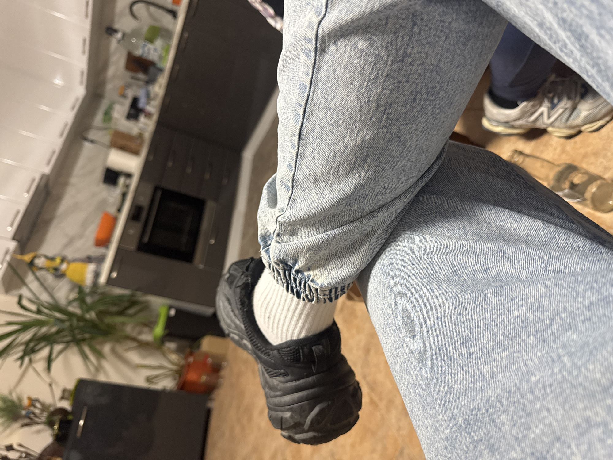 The image depicts a seated individual's legs, crossed at the ankles, within an indoor residential setting identified as a kitchen or combined kitchen/living area in Pleven, Bulgaria. The individual wears light-wash blue denim jeans, white ribbed socks, and a bulky, dark-colored athletic shoe on the left foot. The right foot, partially visible, wears a lighter-colored athletic sneaker over a dark garment. A clear glass bottle lies horizontally on the light-colored floor in the foreground.
The background, largely out of focus, reveals a modern kitchen area with dark grey lower cabinets and white upper wall cabinets. A built-in oven is integrated into the lower cabinetry. A white countertop holds various items, including a black coffee maker, a tall clear bottle, a black faucet, and an orange bowl. To the left, a dark refrigerator or cabinet stands beside several potted plants, one in an orange pot, and a yellow decorative figurine. The room is illuminated by ambient artificial light.