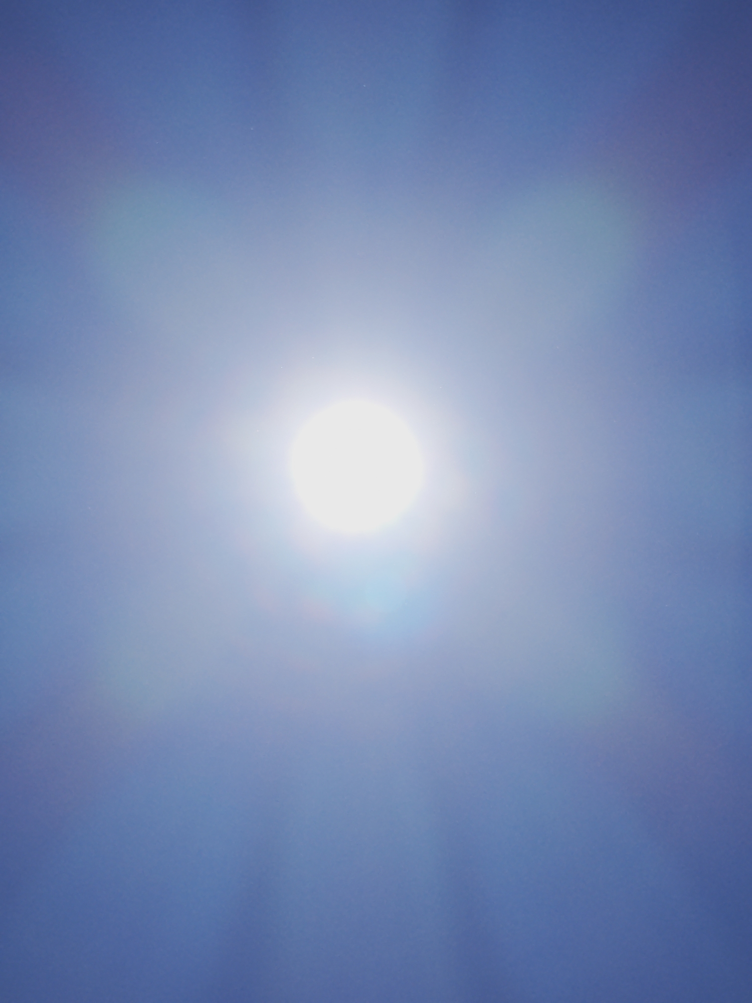 A bright, luminous sun is visible in the center of the image, against a clear blue sky. The sun is slightly out of focus, creating a soft glow and lens flare effects that radiate outwards. The sky transitions from a lighter blue near the sun to a deeper blue at the edges of the frame. The image appears to be taken from an outdoor location in Izgrev, Bulgaria, likely on a clear day. There are no people or specific objects discernible in the scene, only the celestial body and the atmosphere.