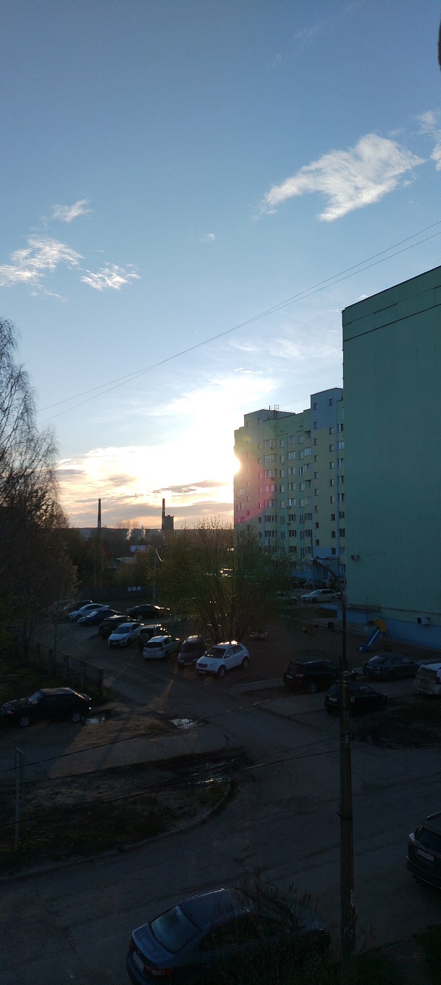 The image shows a cityscape during sunrise or sunset. The sky is blue with scattered clouds, and the sun is positioned behind a multi-story building, creating a bright glare. The building is light green with numerous windows. In the distance, there are two industrial chimneys.
Below the building is a parking lot with various cars parked. A playground area is also visible with a slide. The foreground is a street or courtyard with some vegetation and a few additional cars. A utility pole stands near the right edge of the image.