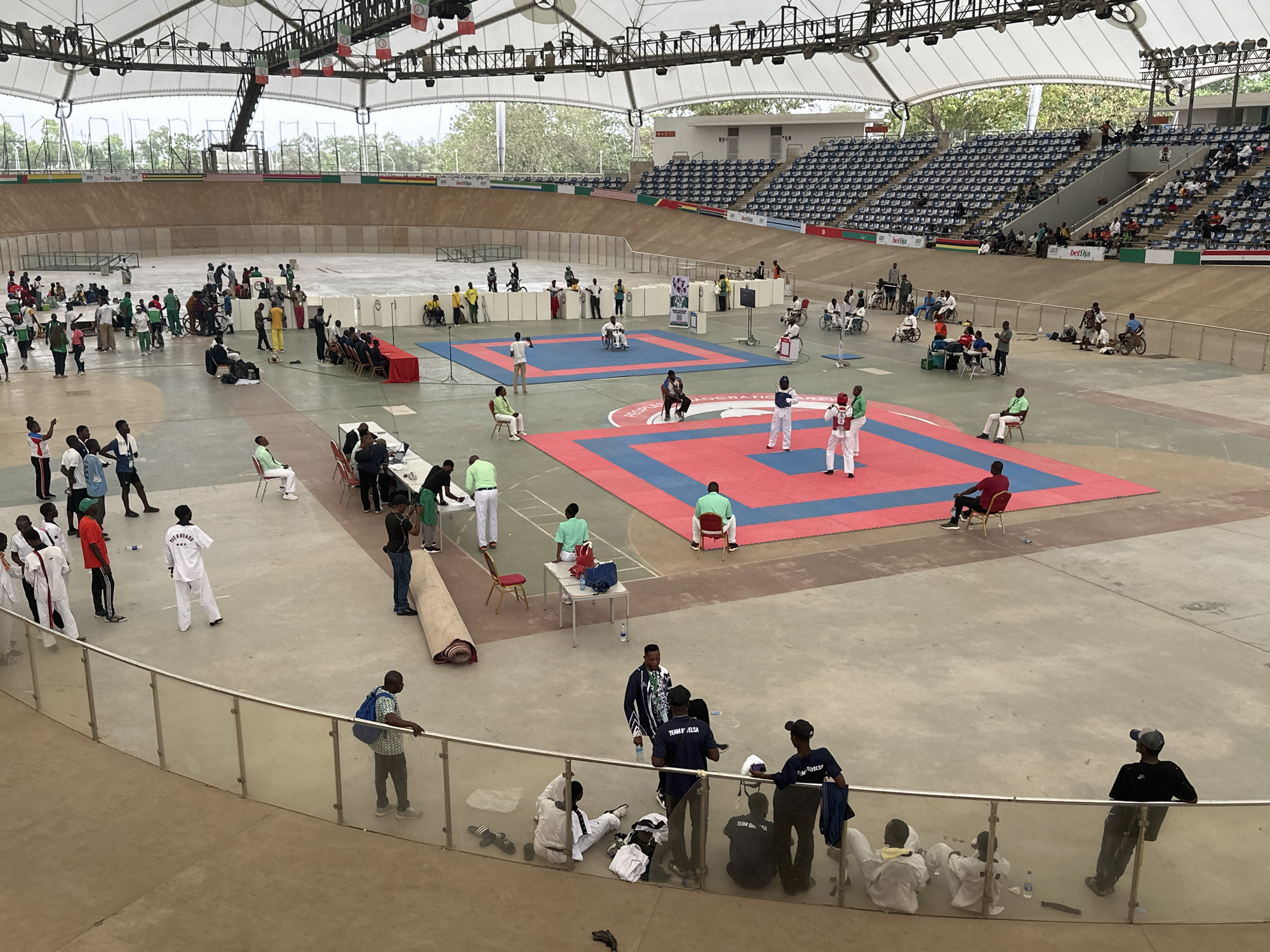 The image depicts an indoor sporting event, possibly a taekwondo competition, taking place in an arena. Two athletes are engaged in a match on a red and blue mat, while other individuals, presumably coaches or officials, are seated nearby. Spectators are visible in the stands. In the foreground, there are people standing and sitting, observing the event. The arena's architecture includes a high ceiling with visible support structures and lighting fixtures. The event seems to be held in the city of Unknown, Unknown. The presence of a track-like structure surrounding the central area suggests the arena can be used for multiple sports.
