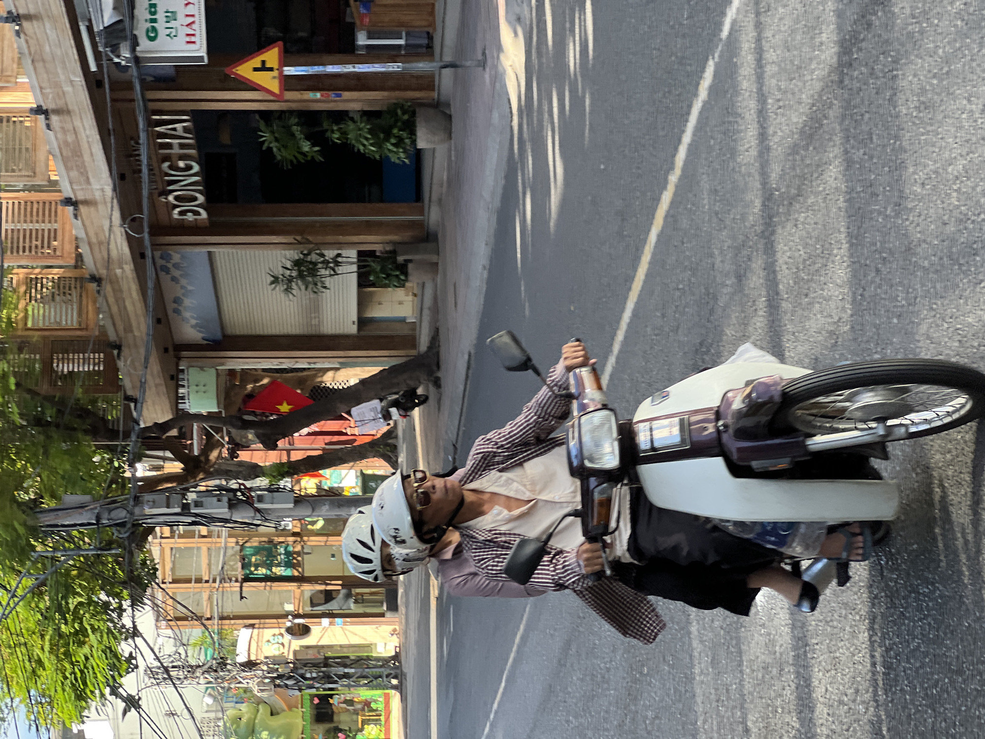 Two individuals are riding a scooter on a street in Nha Trang, Vietnam. The person in front, wearing a white helmet and sunglasses, is operating the scooter. They are dressed in a light-colored shirt with a striped pattern and dark pants. The person behind them, also wearing a white helmet, is wearing a pink shirt and dark pants. The scooter is white and maroon. In the background, buildings with wooden facades and signs are visible, including one that reads "ĐÔNG HAI". A yellow triangular traffic sign with a black T-shaped symbol is also present. Trees with green foliage line the street, and power lines are strung overhead. The road surface is asphalt with a yellow line marking. Shadows from the trees are cast across the road.