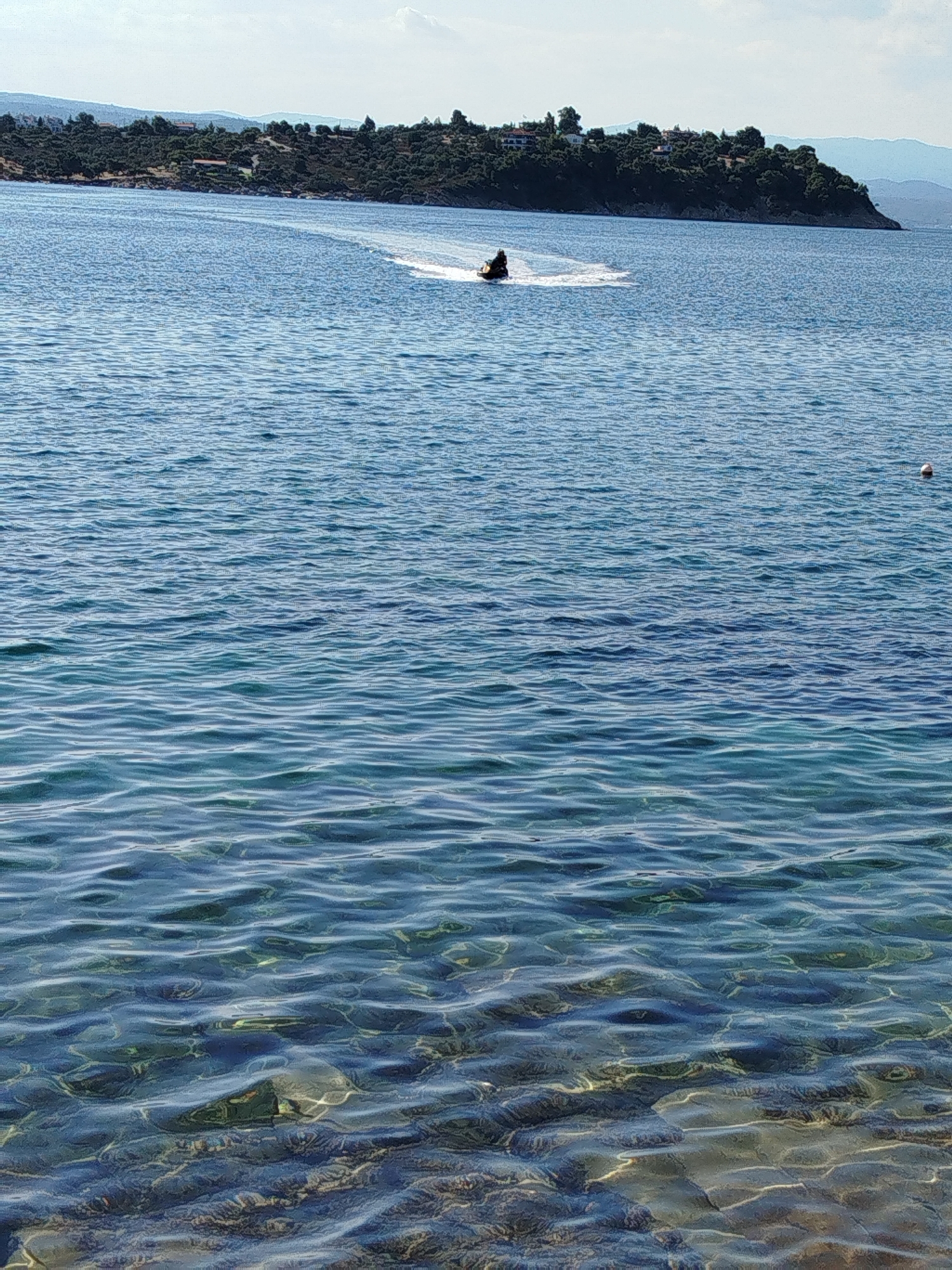 In the Sithonia Municipal Unit, Greece, this image captures a serene coastal scene. The foreground features clear, shallow turquoise water with visible rocky seabed illuminated by sunlight. The midground is dominated by the vast expanse of the Aegean Sea, with gentle waves rippling across its surface. A lone jet ski rider is visible in the distance, leaving a white wake as they glide across the water. The background showcases a lush, green coastline with a cluster of buildings nestled amongst trees, suggesting a residential area. Rolling hills and distant mountains form the horizon under a bright, partially cloudy sky. A small, red and white buoy floats on the water to the right of the frame, adding a subtle detail to the seascape.