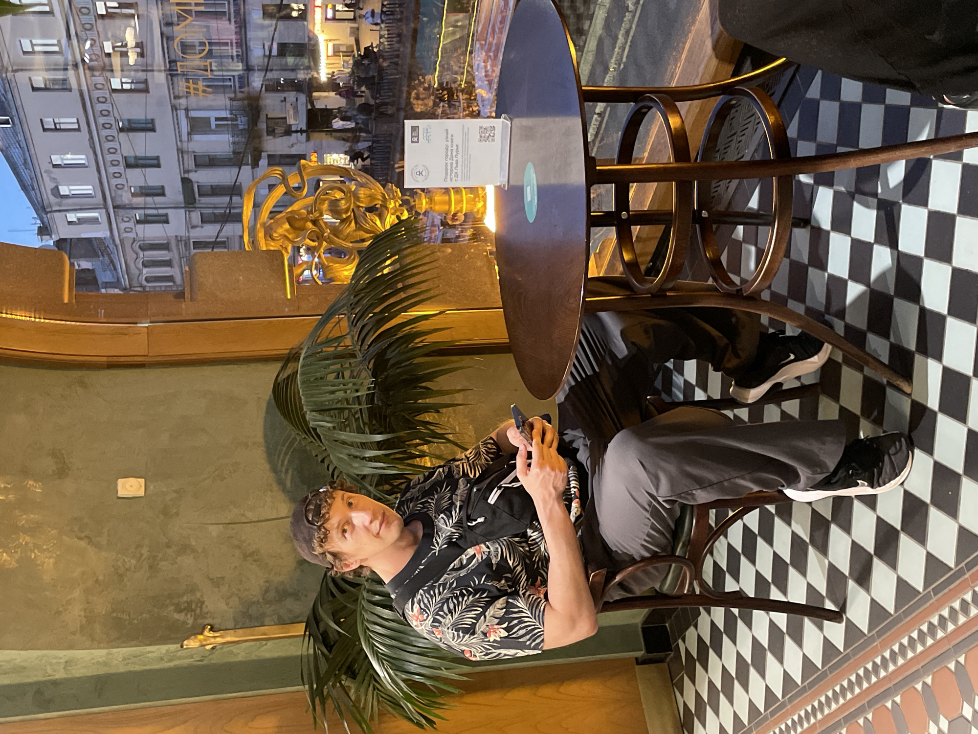 A man with curly brown hair, wearing a dark baseball cap backward, a black tropical-patterned t-shirt, gray pants, and black sneakers, sits in a dark wooden chair at a round, dark wooden table. He has a black fanny pack across his chest and holds a smartphone in both hands, looking towards the camera with a slight smile. Behind him is a large green palm plant against a dark green textured wall, with a small white electrical outlet or sensor visible on the wall.
On the table, a white sign in a clear stand displays Russian text, including "Подарок городу: узнай историю Дома книги и Дома Зингера" (A gift to the city: learn the history of the House of Books and the Singer House), along with a QR code. The floor is covered in a black and white checkerboard tile pattern, with a decorative border of reddish-brown and lighter tiles.
Through a large window, an evening view of a city street in Saint Petersburg, Russia, is visible. The buildings across the street exhibit classical Russian architecture, with multiple windows and balconies, under a twilight sky. Streetlights and ground-floor shop lights are illuminated, and people can be seen walking on the street. A large, ornate golden sculpture or decorative element is visible near the window, and a partial hashtag "#ДОМКН" (likely referring to "Дом книги" or House of Books) is visible on the window glass. The presence of the sign explicitly mentioning "Дома Зингера" (Singer House) identifies the location as being at or in close proximity to the renowned Singer House (Dom Knigi) at Nevsky Prospect, 28, in Saint Petersburg.