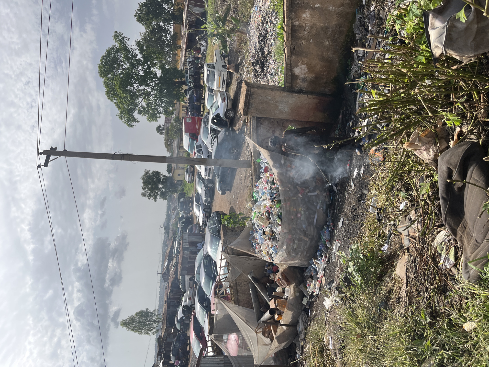 The image depicts an outdoor scene in Jos, Nigeria, likely a junkyard or a waste processing area, with a heavy accumulation of discarded items. In the foreground, a large pile of plastic bottles, mostly clear and some colored, are contained within a large, translucent bag or tarp. Adjacent to this pile, there is a smaller collection of various plastic containers and what appears to be discarded pipes. Some of this material is smoldering, indicated by plumes of smoke rising from the pile and the dark, charred ground. A person, partially obscured by smoke and vegetation, can be seen near the fire, potentially tending to it or salvaging materials.
Behind the immediate waste piles, a long line of abandoned or dismantled vehicles is visible, ranging from cars to what looks like a red truck. These vehicles are parked closely together, suggesting a state of disrepair or storage. Beyond the vehicles, there are rudimentary structures and buildings, indicating a densely populated or developed area adjacent to the junkyard. Lush green trees are scattered throughout the scene, some reaching considerable heights, contrasting with the otherwise desolate environment. The sky is overcast with scattered clouds, suggesting either early morning or a cloudy day, with no strong indication of the sun's position. The overall atmosphere conveyed is one of a working junkyard, possibly involved in recycling or disposal, with an element of environmental concern due to the burning waste. No visible text is present in the image.