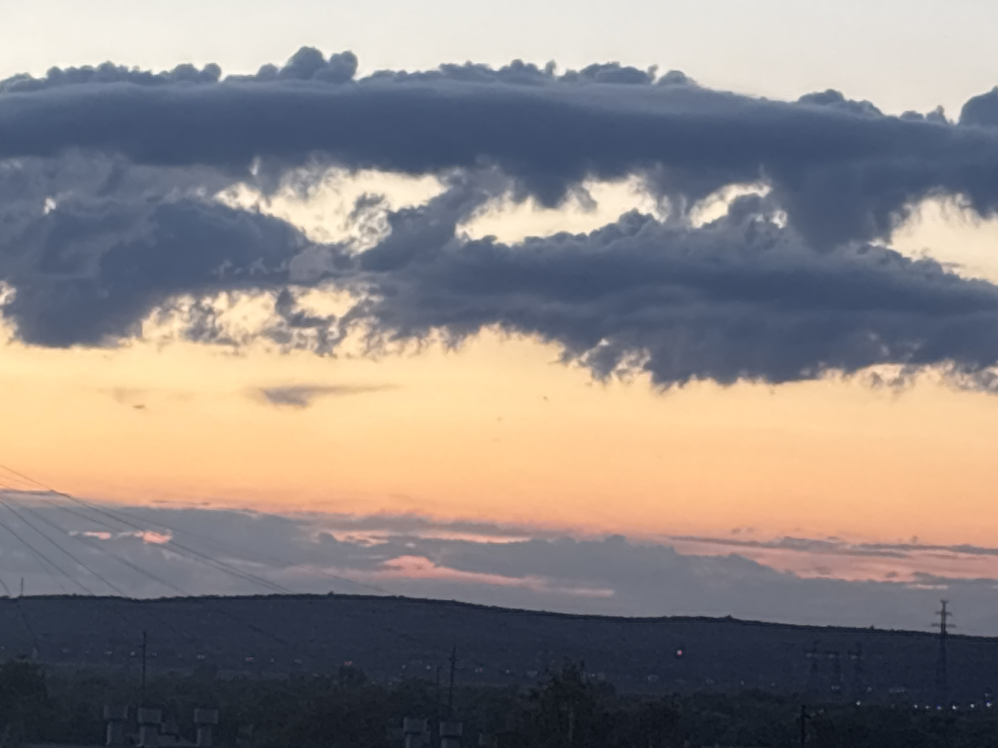 The image captures a serene twilight landscape in Chaikovsky, Russia, during what appears to be sunset or dusk. The sky dominates the frame, showcasing a dramatic interplay of light and shadow.
In the upper two-thirds of the image, heavy, dark grey clouds are prominent, forming a distinct horizontal band. These clouds are thick and layered, with lighter, warm-toned sky peeking through irregular openings, creating bright, almost glowing patches within the cloud mass. Below this dark cloud layer, the sky transitions to vibrant hues of orange and yellow near the horizon, gradually fading to a lighter, soft blue-grey at the very top of the frame. A lighter, more diffuse band of clouds, tinged with subtle pinks and purples from the setting sun, stretches across the mid-sky, partially obscuring the distant landscape.
Below the colorful sky, the distant terrain appears as a dark, undulating silhouette of hills or mountains. Faint, pinprick lights, possibly from a distant settlement or industrial area, are visible along the horizon line. In the mid-ground and foreground, dark, silhouetted structures, including what appear to be power lines and utility poles or towers, are visible against the fading light. The overall mood of the image is peaceful and contemplative, highlighting the natural beauty of the sky at dusk. There is no visible text in the image.