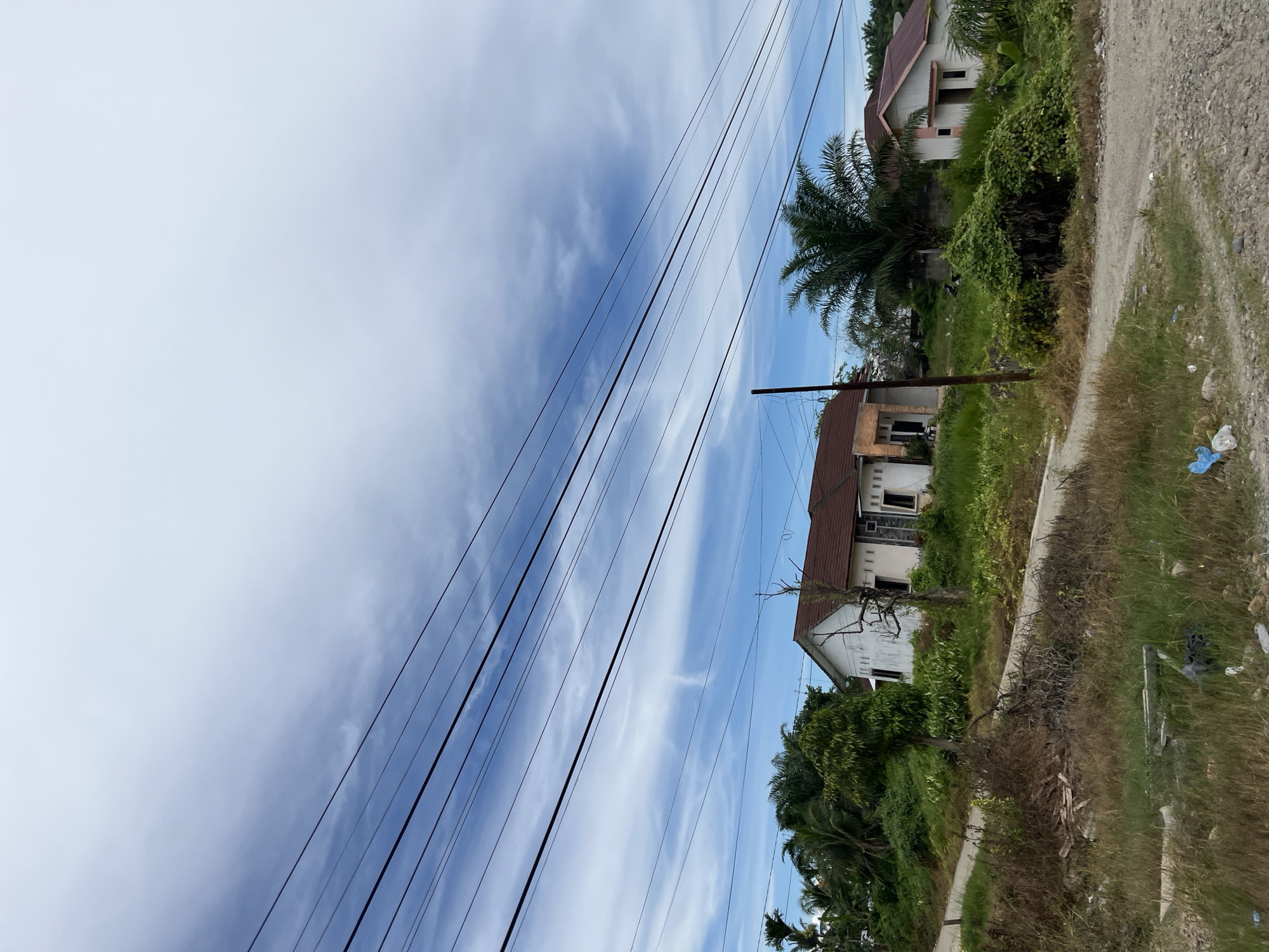 A low-angle shot depicts a cloudy sky over a rural landscape in Seunebobaru, Indonesia. Multiple dark power lines stretch across the upper portion of the frame, dividing the sky and obscuring some of the distant view. In the middle ground, a cluster of houses, some appearing under construction or in disrepair, are nestled amongst lush green vegetation and palm trees. A dirt road winds through the scene, with dry grass and scattered debris in the foreground. The overall impression is one of a developing or somewhat neglected rural settlement.