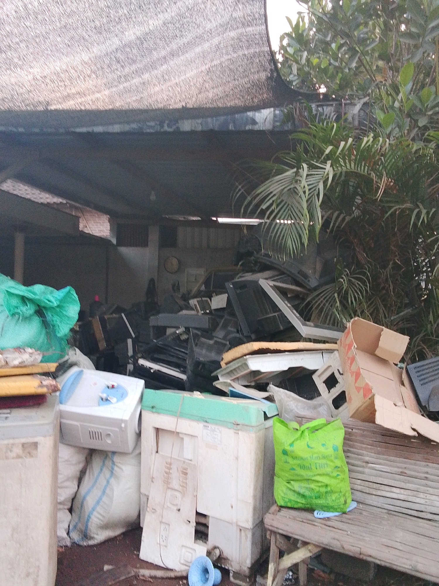 The image depicts an outdoor storage area filled with discarded electronics and appliances, likely in a recycling or waste management facility in Bulurejo, Indonesia. A large, dark-colored shade structure covers a significant portion of the area, providing some protection from the elements. To the right, lush green foliage from trees and plants encroaches on the scene.
Dominating the foreground are various electronic items. A white appliance, possibly a water dispenser, sits atop large white sacks with blue stripes. To its left, a cream-colored washing machine or similar appliance is partially visible. To the right of the white dispenser, another appliance with a mint-green top and a white front panel is stacked with what appears to be electronic components. A bright green plastic bag, printed with text, rests on a weathered wooden bench in front of this appliance.
Further back, piles of electronic waste are visible, including what appears to be old computer monitors, television screens, and other electronic components. A damaged cardboard box with red and white markings is also present. The ground is covered with debris and a reddish-brown surface, suggesting it's an unpaved area. In the background, glimpses of a metal roof and some brickwork indicate the presence of a building or structure. The overall scene conveys a sense of disarray and the accumulation of outdated or broken technology. There are no visible people in the image.