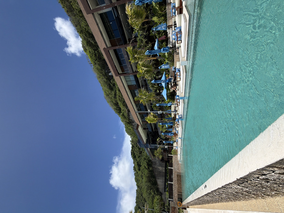 A bright blue swimming pool occupies the right side of the frame, bordered by a white coping. Beyond the pool, a building nestled into a lush, green hillside is visible. Numerous blue umbrellas and lounge chairs line the pool area. The sky, a vibrant blue, is sparsely populated with white clouds. The composition is shot from a slightly low angle, emphasizing the height of the building and the expanse of the sky.