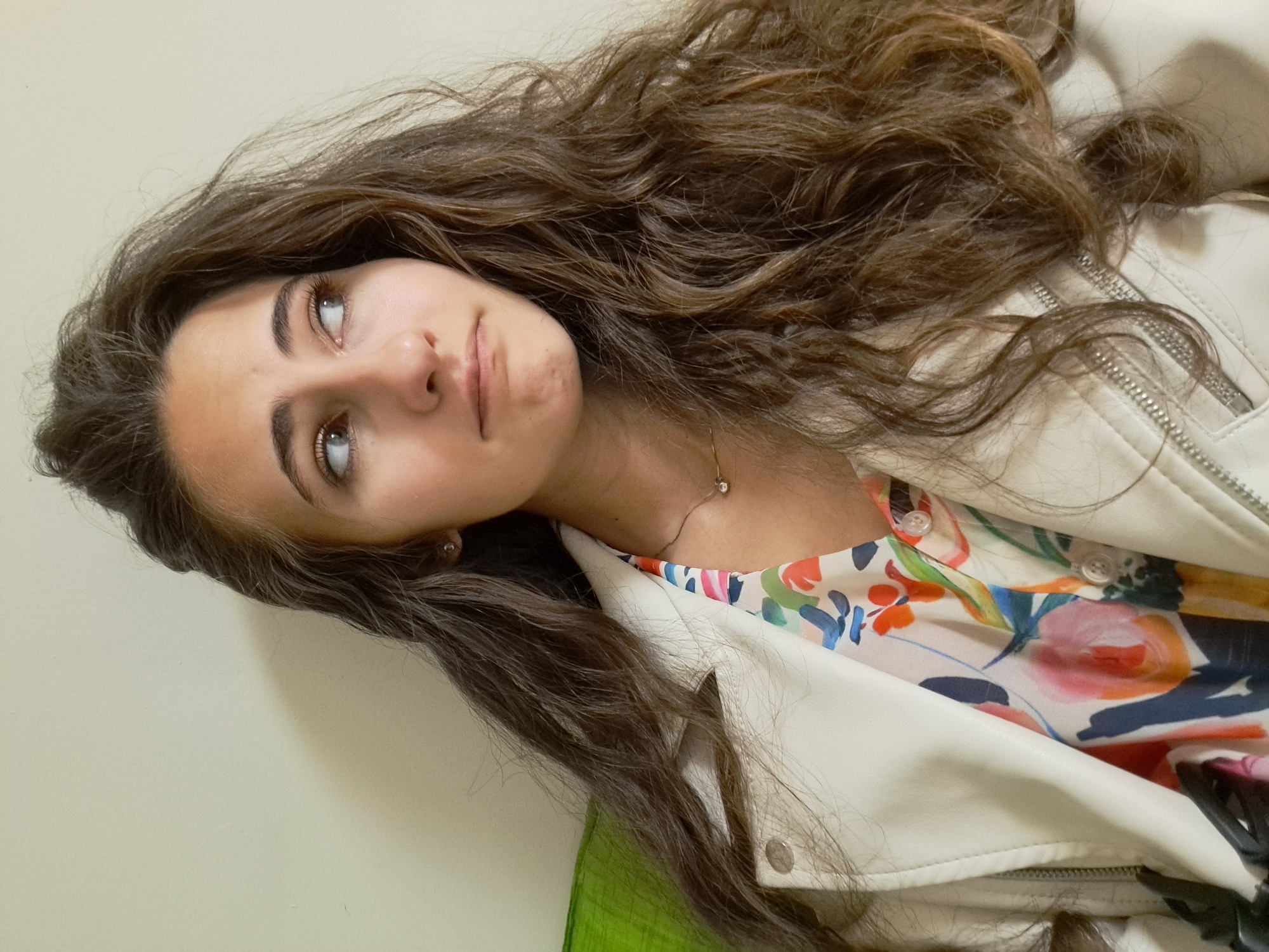 A young woman with long, wavy brown hair is posed at an angle against a light-colored wall. She is wearing a floral-patterned top underneath a white leather jacket. A gold necklace is visible around her neck. Her gaze is directed upwards and slightly to the left. The location is not identifiable from the image.