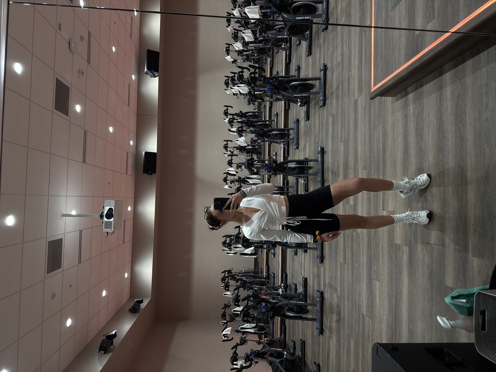 A photograph shows a young woman taking a selfie in an indoor cycling studio. She stands in the aisle between rows of stationary bikes, facing a mirror. The woman has dark hair pulled back and wears a white, long-sleeved shirt and black shorts. She holds a black smartphone up to her face, presumably to capture her reflection.
The cycling studio is well-lit with a high ceiling covered in recessed lighting. Additional lighting fixtures are mounted on the walls. A projector hangs from the ceiling on the left side of the image. The floor is made of a dark wood-like material. There are rows of stationary bikes on either side of the aisle, all facing forward. The location is in the city of Irvine, United States of America, based on the "FIVE LINE" decal on each bike.