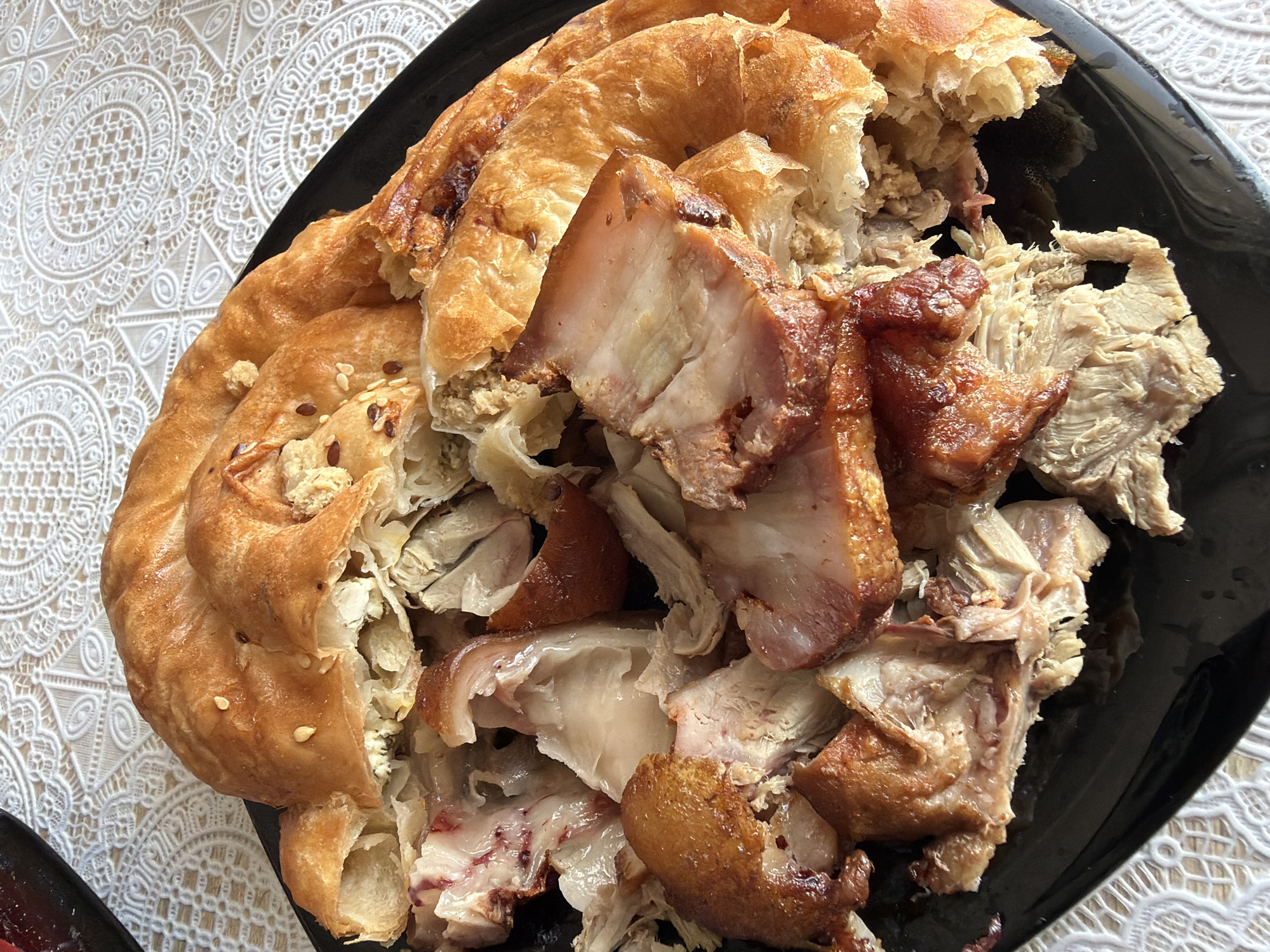 A close-up, high-angle shot captures a black plate filled with a portion of a golden-brown, spiral-shaped pastry, likely a savory pie. The pastry is torn open, revealing a generous filling of shredded meat and possibly some cheese or vegetables. Several pieces of cooked meat, some with visible fat and crispy skin, are scattered over the pastry. The plate rests on a white tablecloth with an intricate lace-like pattern. The setting is a domestic environment, possibly a kitchen or dining room in Strășeni, Moldova. No people are visible in the image.