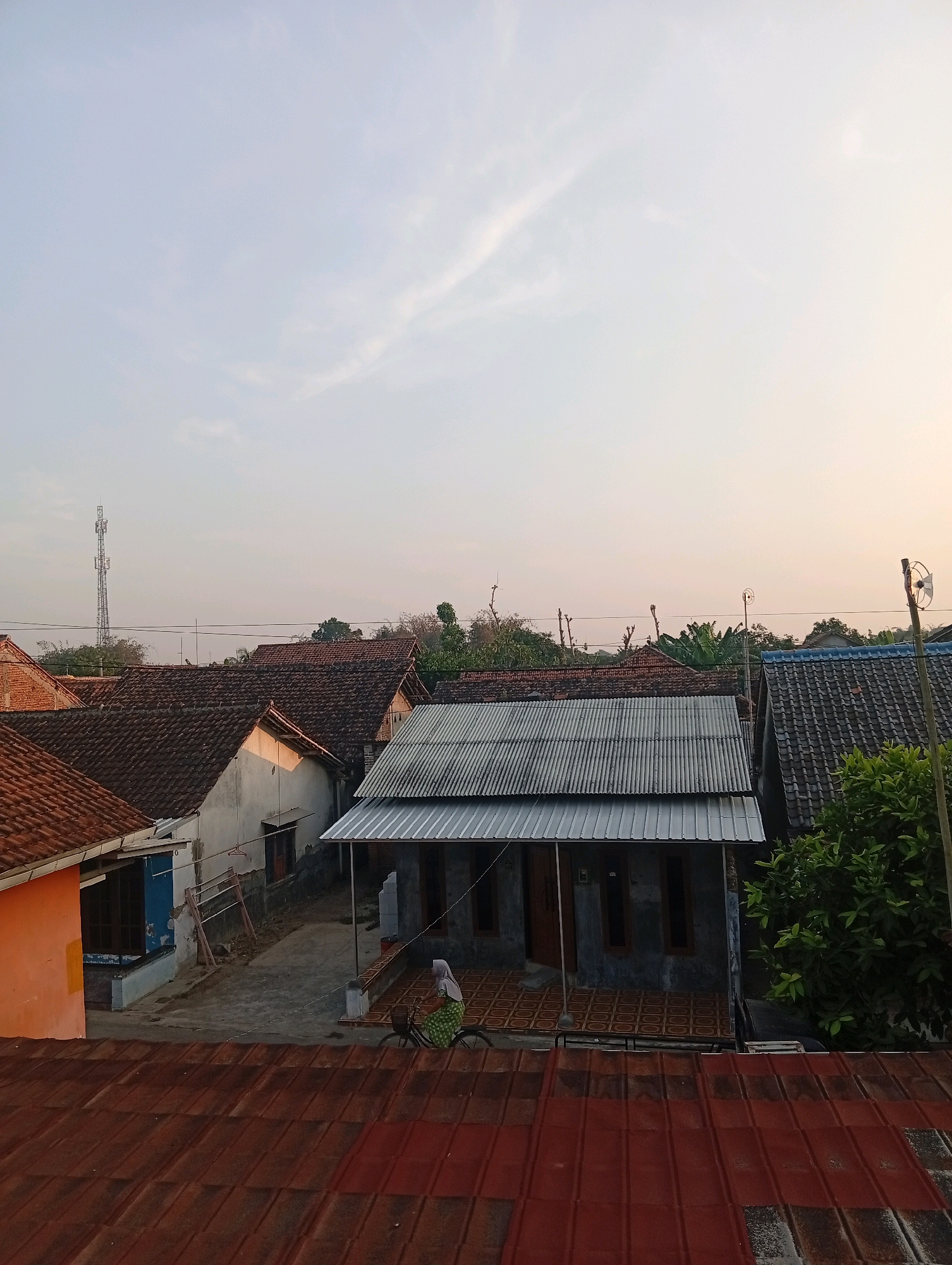 This image captures a residential scene in Kebandingan, Indonesia, likely at dawn or dusk given the warm, soft lighting and pale sky. The foreground is dominated by the reddish-brown tiled roofs of several houses. Beyond these, a street or pathway is visible, with a person riding a bicycle. This individual is wearing a headscarf and a patterned dress. They are cycling past a modern-looking house with a gray, corrugated metal roof and darker walls, featuring multiple windows and a wooden door. Adjacent to this house, another building with a white facade and a traditional red-tiled roof is visible. Further in the background, more rooftops and greenery are present. A tall telecommunications tower stands against the horizon on the left side of the image, indicating a developed area. The overall impression is of a quiet, everyday moment in a rural or suburban Indonesian neighborhood.