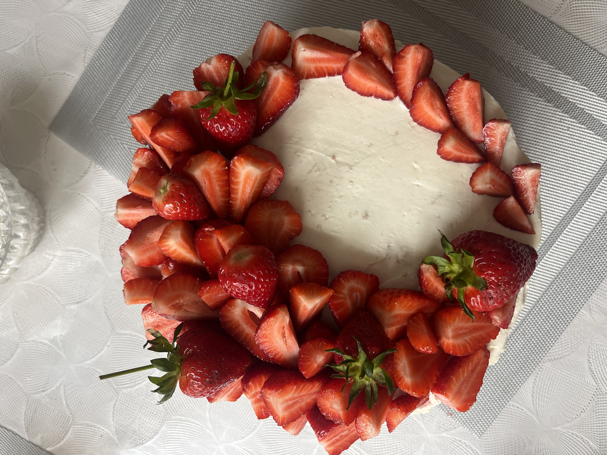 The image shows a strawberry cake on a table. The cake has a white frosting and is decorated with whole and sliced strawberries. The strawberries are arranged around the edge of the cake, leaving the center of the frosting visible. The cake is sitting on a white tablecloth with a textured pattern. The setting appears to be indoors. There is no information in the image to place the location in Kaspiysk, Russia or to identify any people, actions, or interactions.